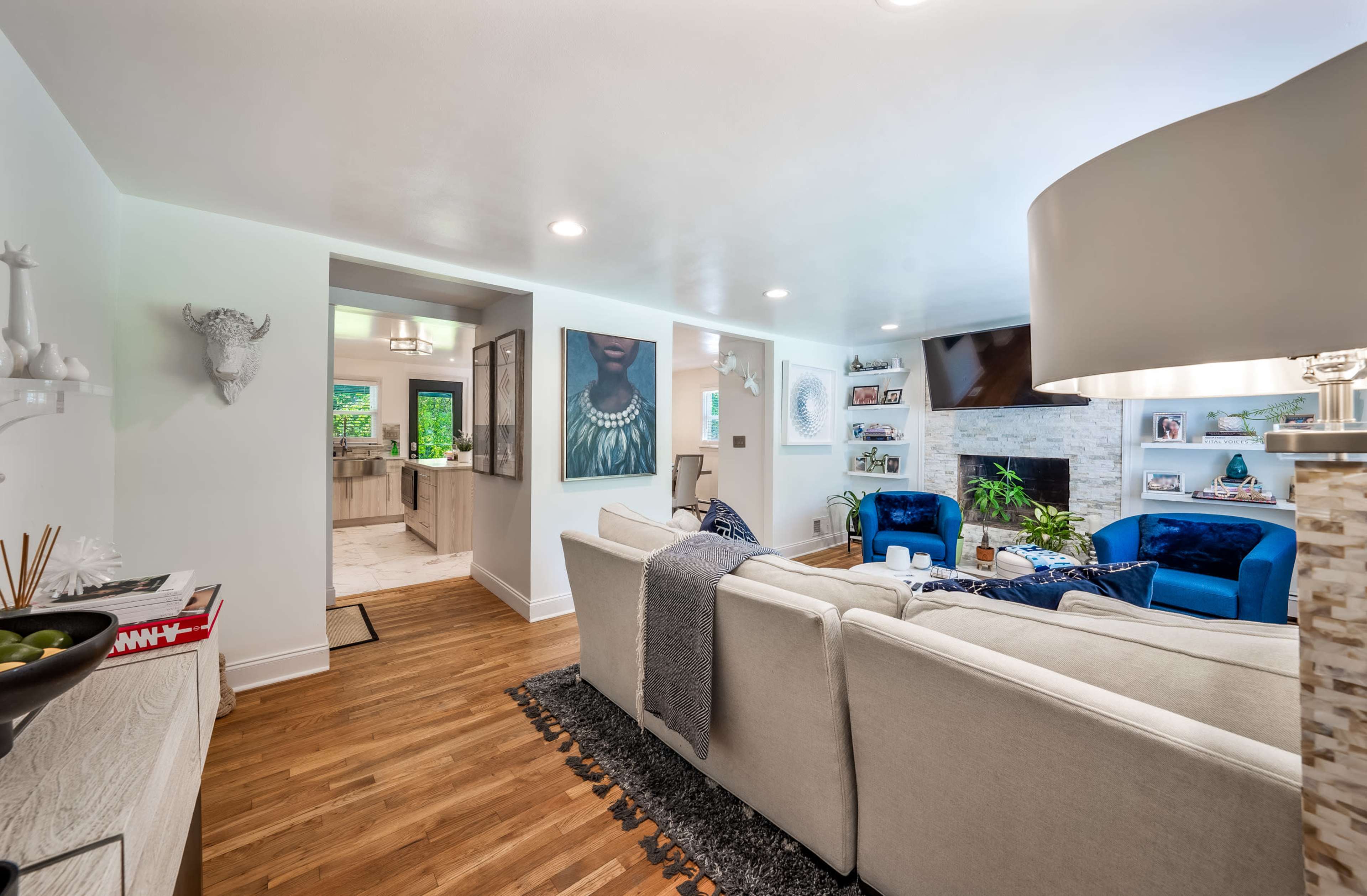 A modern living room with a beige sectional couch, two blue accent chairs, a television above a stone fireplace, and two artworks on the walls, leading to a kitchen area.