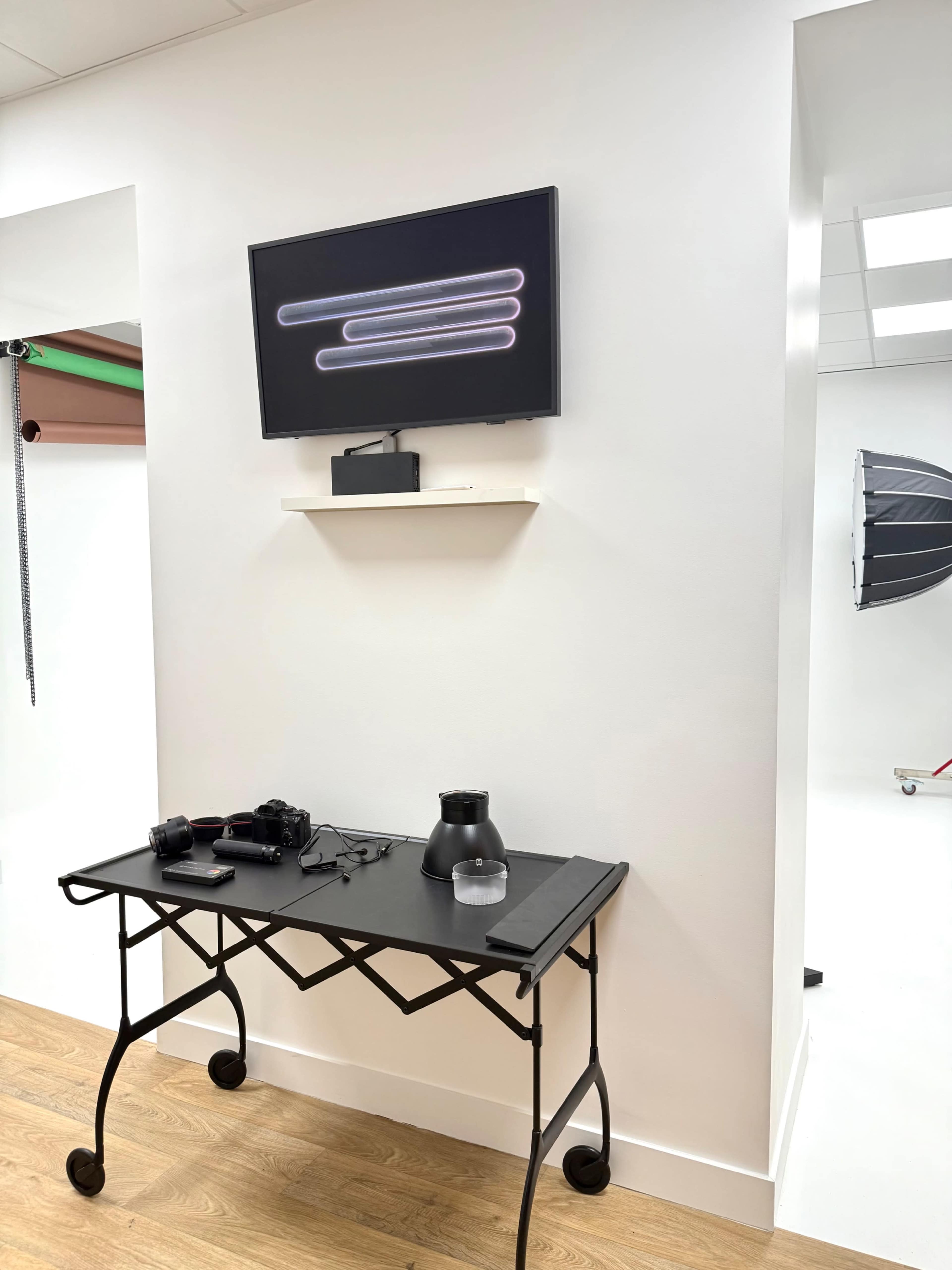 A minimalist studio space features a mounted television above a small black table that holds photography equipment and a vase.
