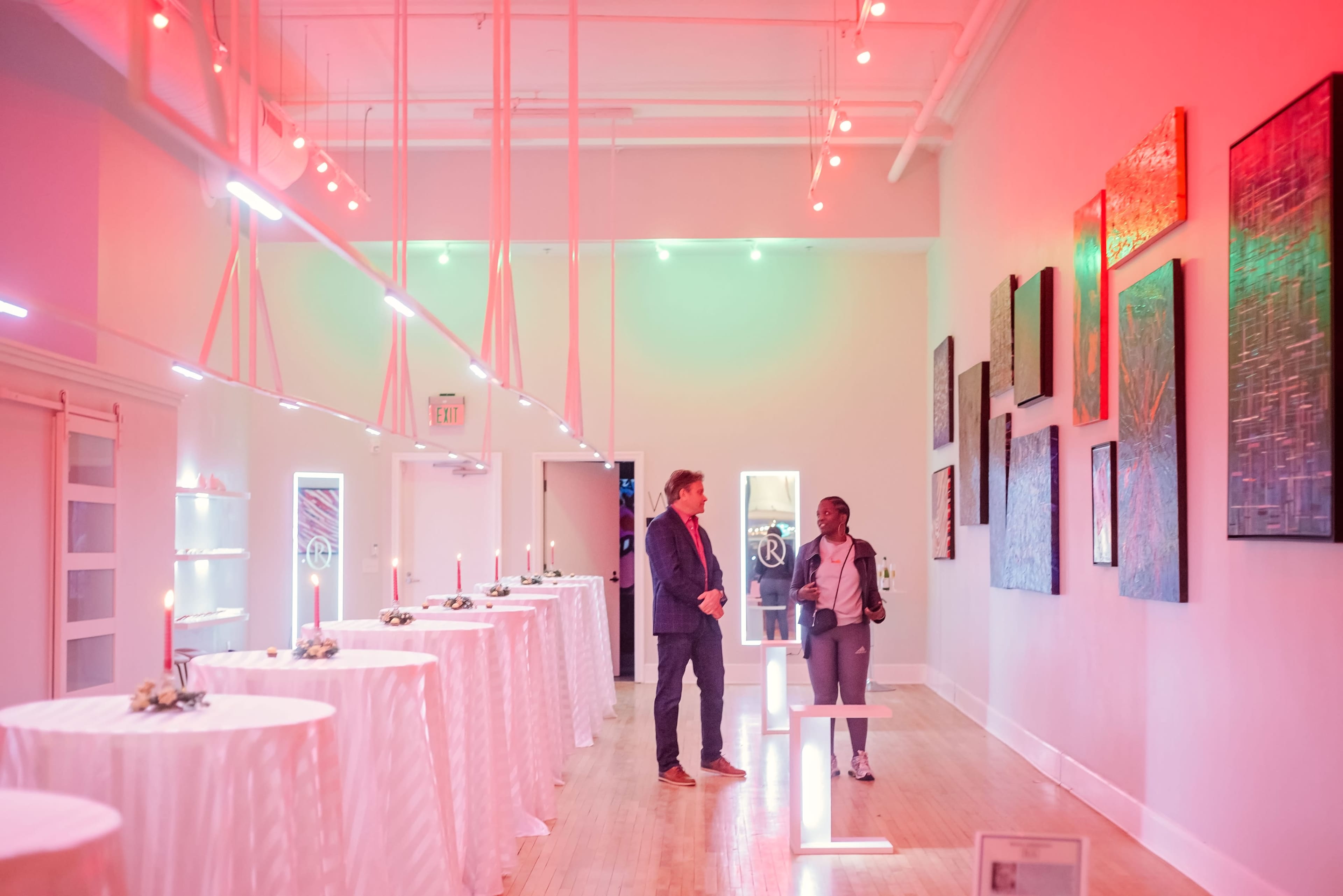 Two individuals converse in a brightly lit art gallery featuring white tables and vibrant wall art.