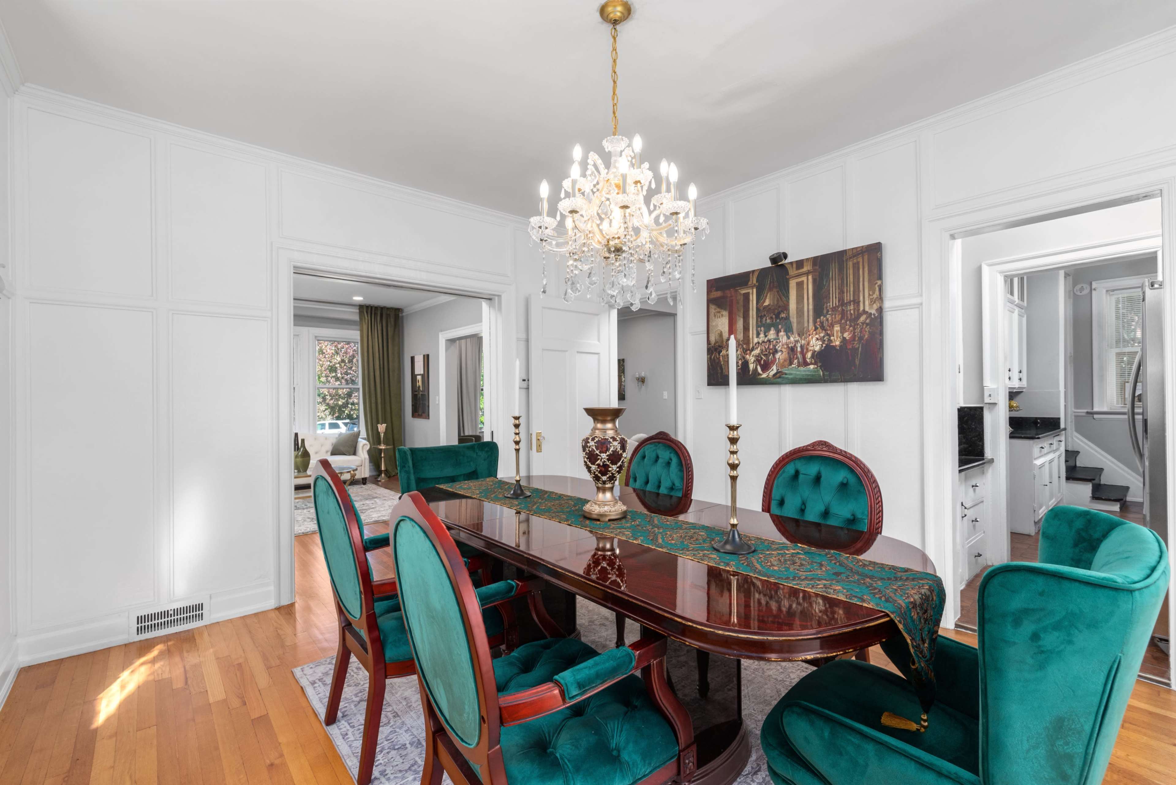 The dining room features a dark wooden table surrounded by six teal upholstered chairs and a crystal chandelier overhead.