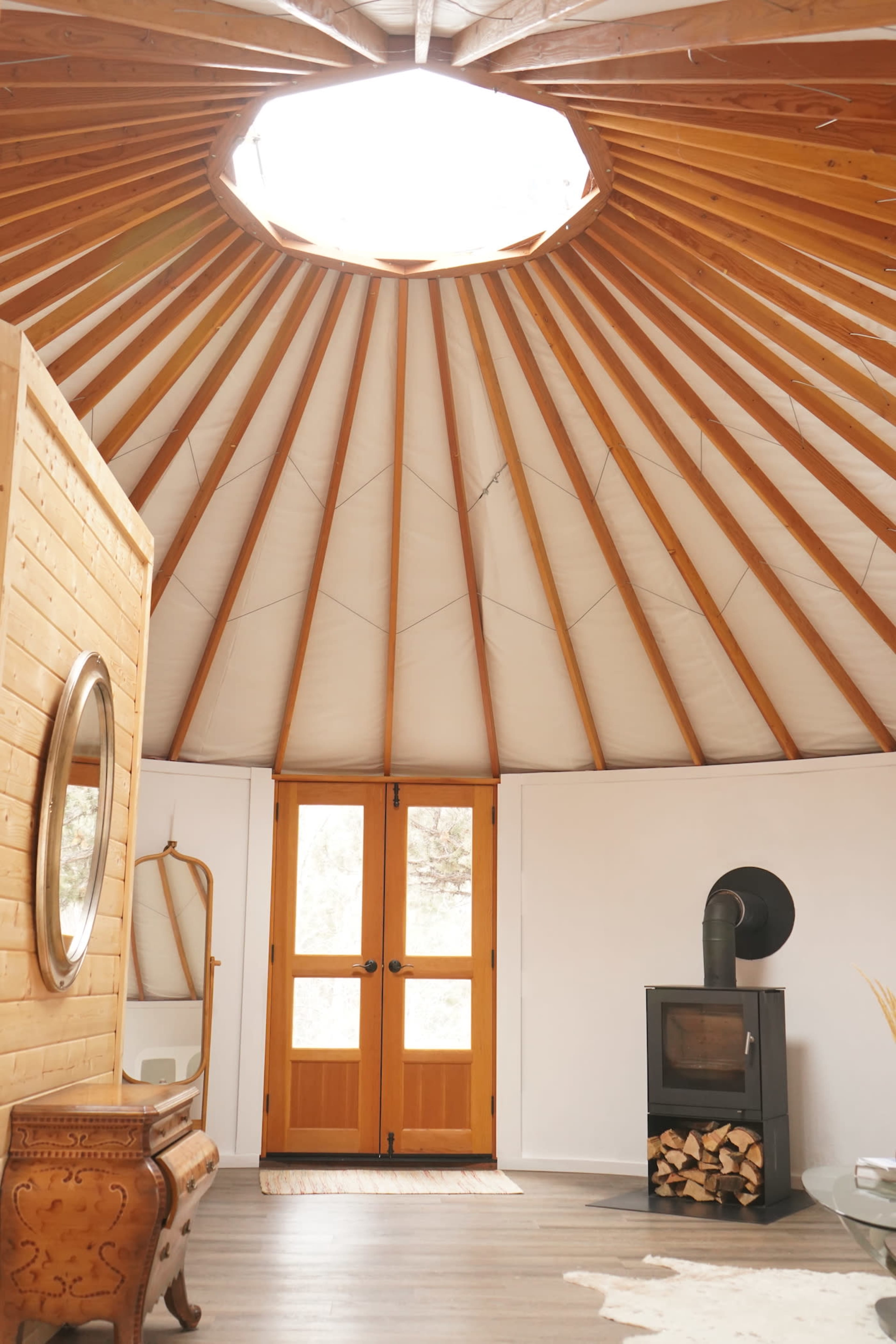 The interior of a yurt features a circular wooden ceiling, a central skylight, a wood stove, and two doors leading outside.