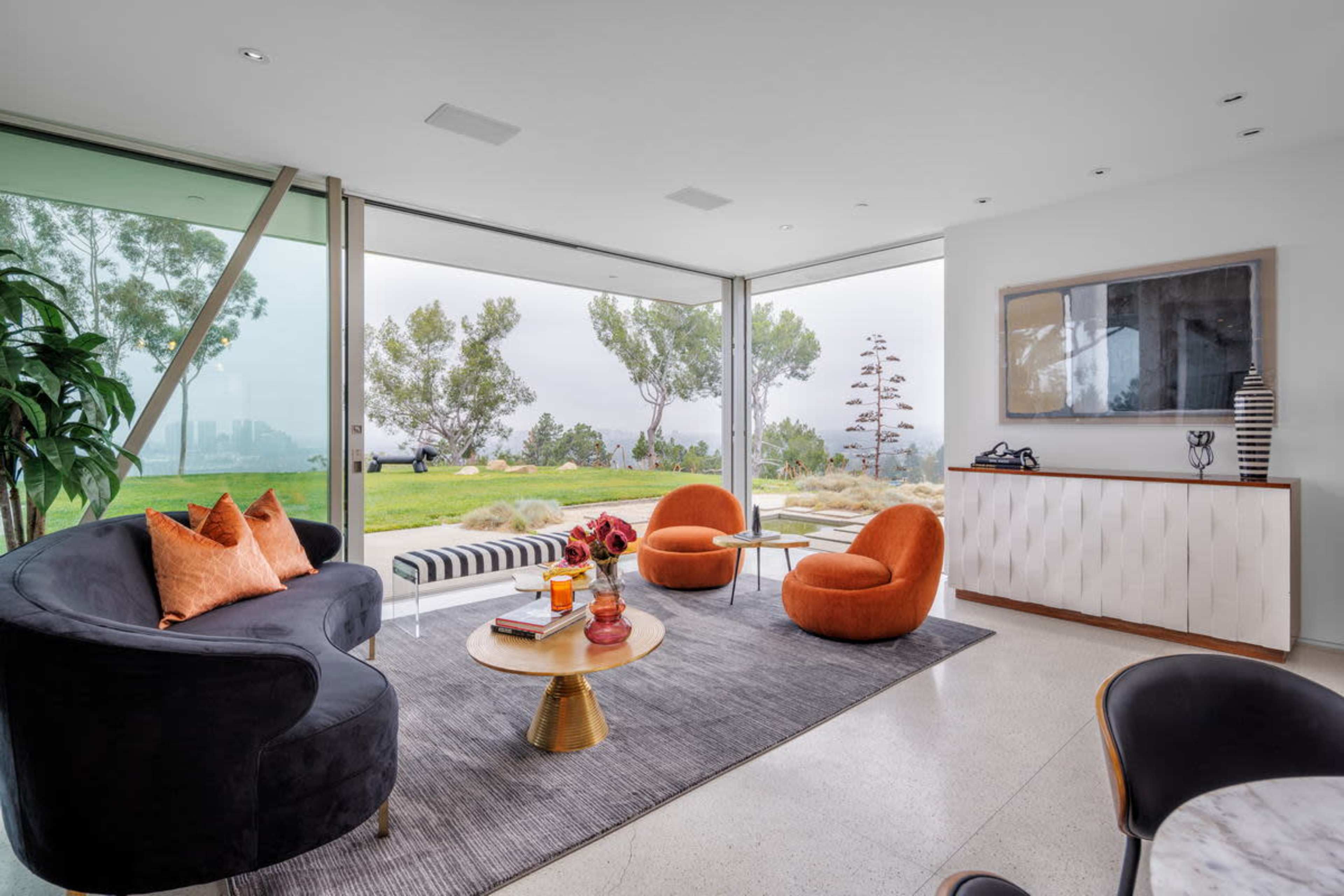 A modern living room features large glass windows, a black sofa, two orange armchairs, a round coffee table, and a white sideboard, overlooking a green landscape.