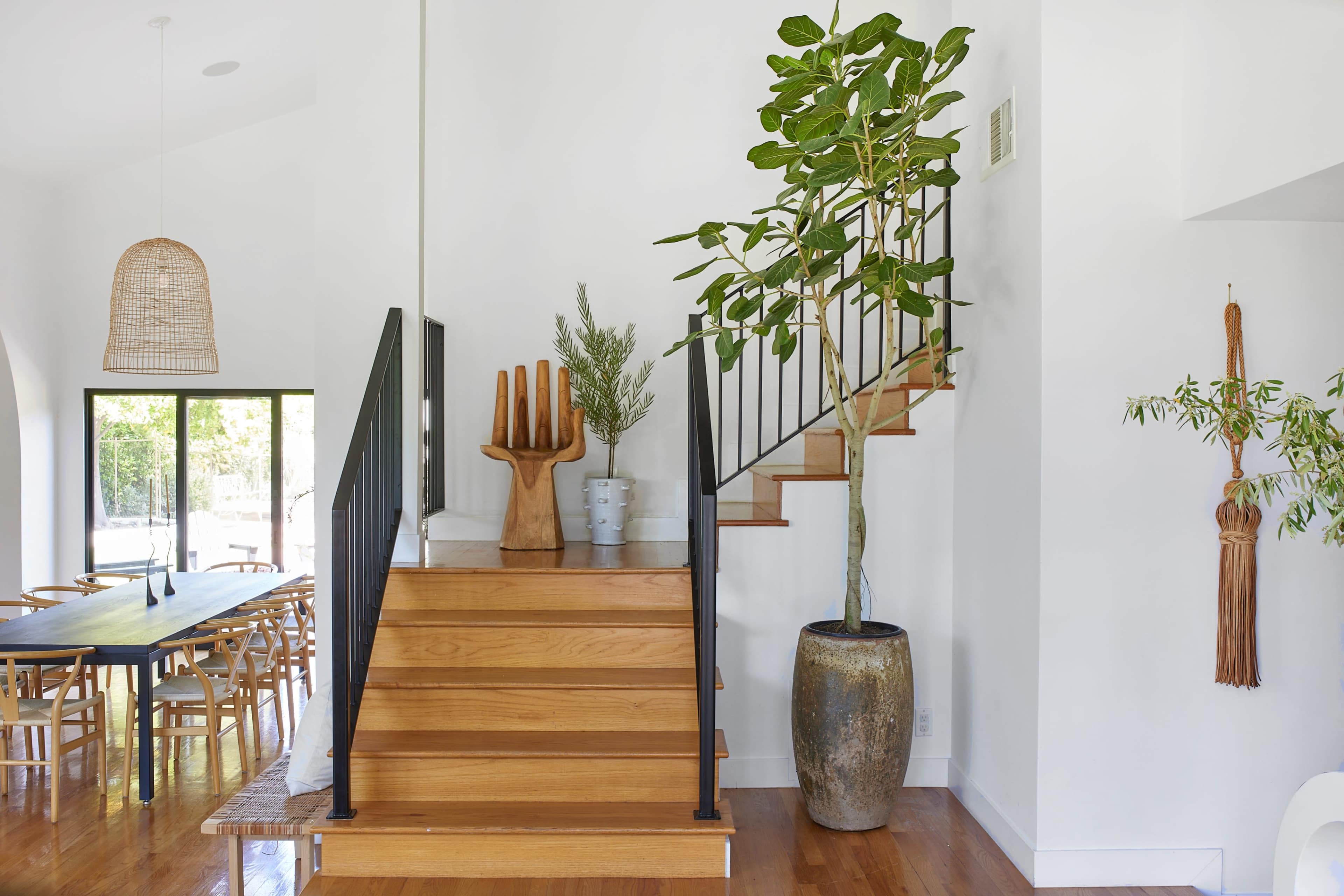 A modern interior with a staircase leading to an upper level, adorned with a large potted plant and wooden decor, alongside a dining area visible through a large window.