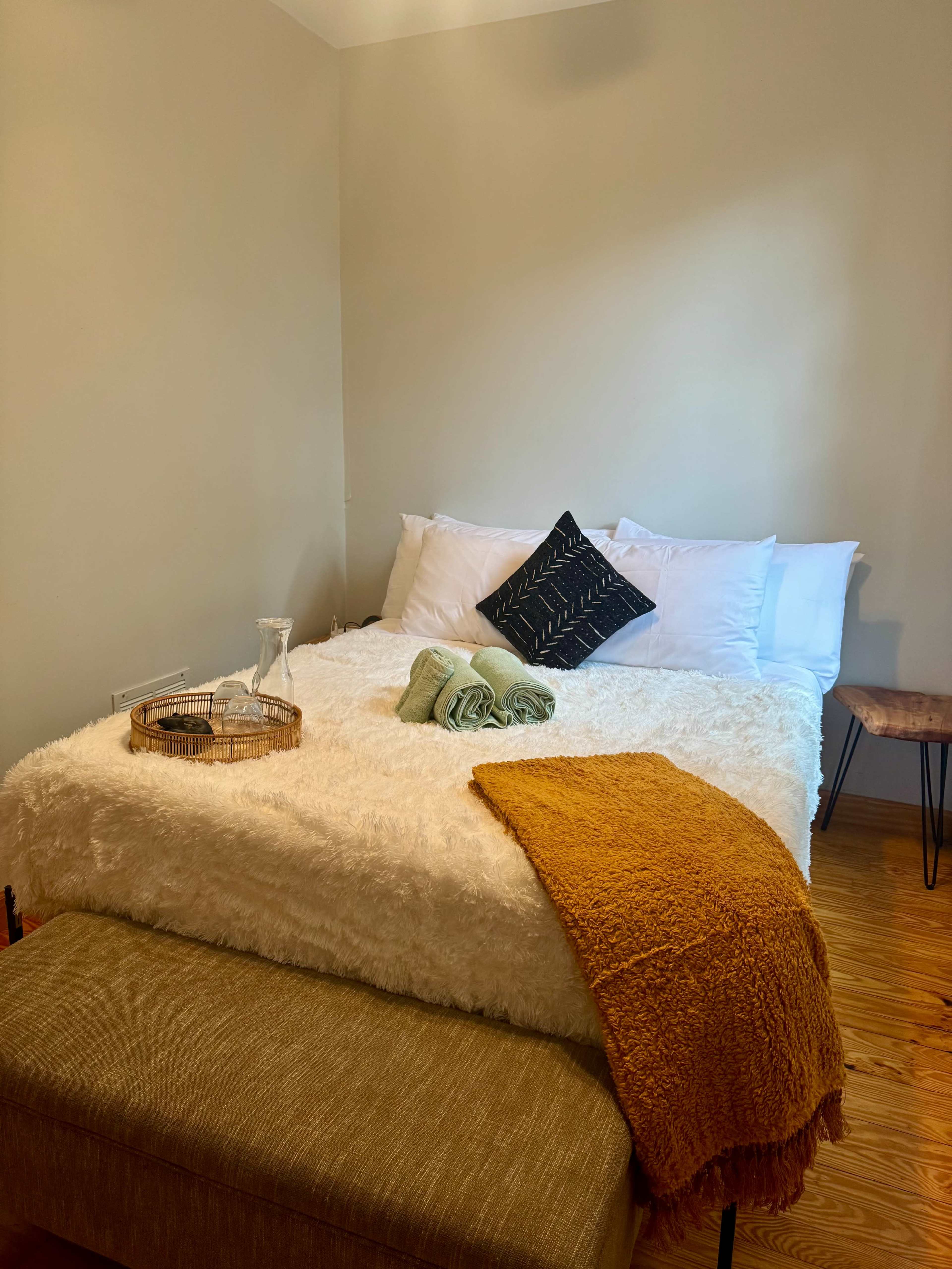 The image shows a cozy bedroom with a neatly made bed, a wooden side table, and a bench at the foot of the bed.