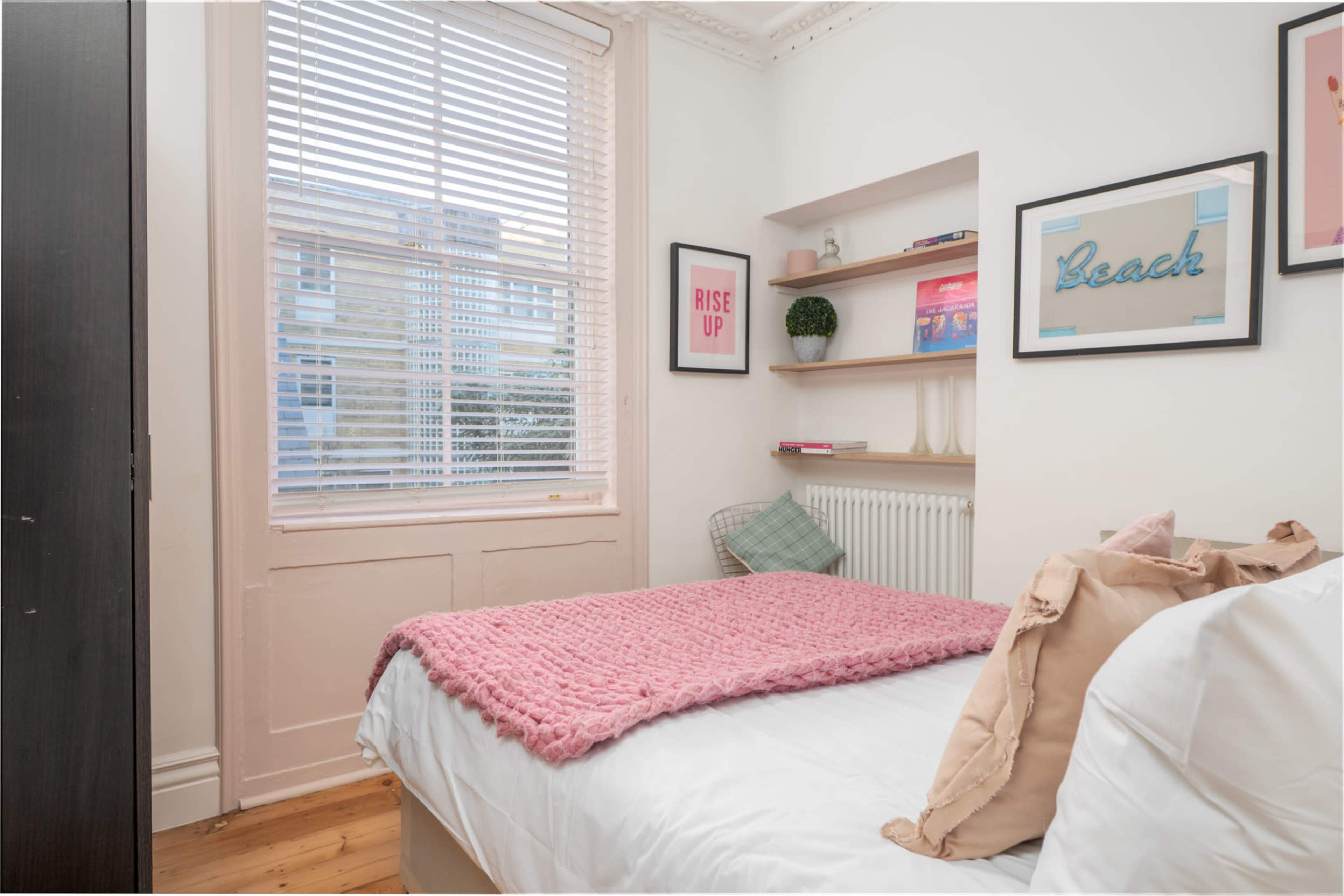 The image shows a small bedroom with a bed covered in a pink blanket, a shelf with decor items, and a window with blinds letting in natural light.
