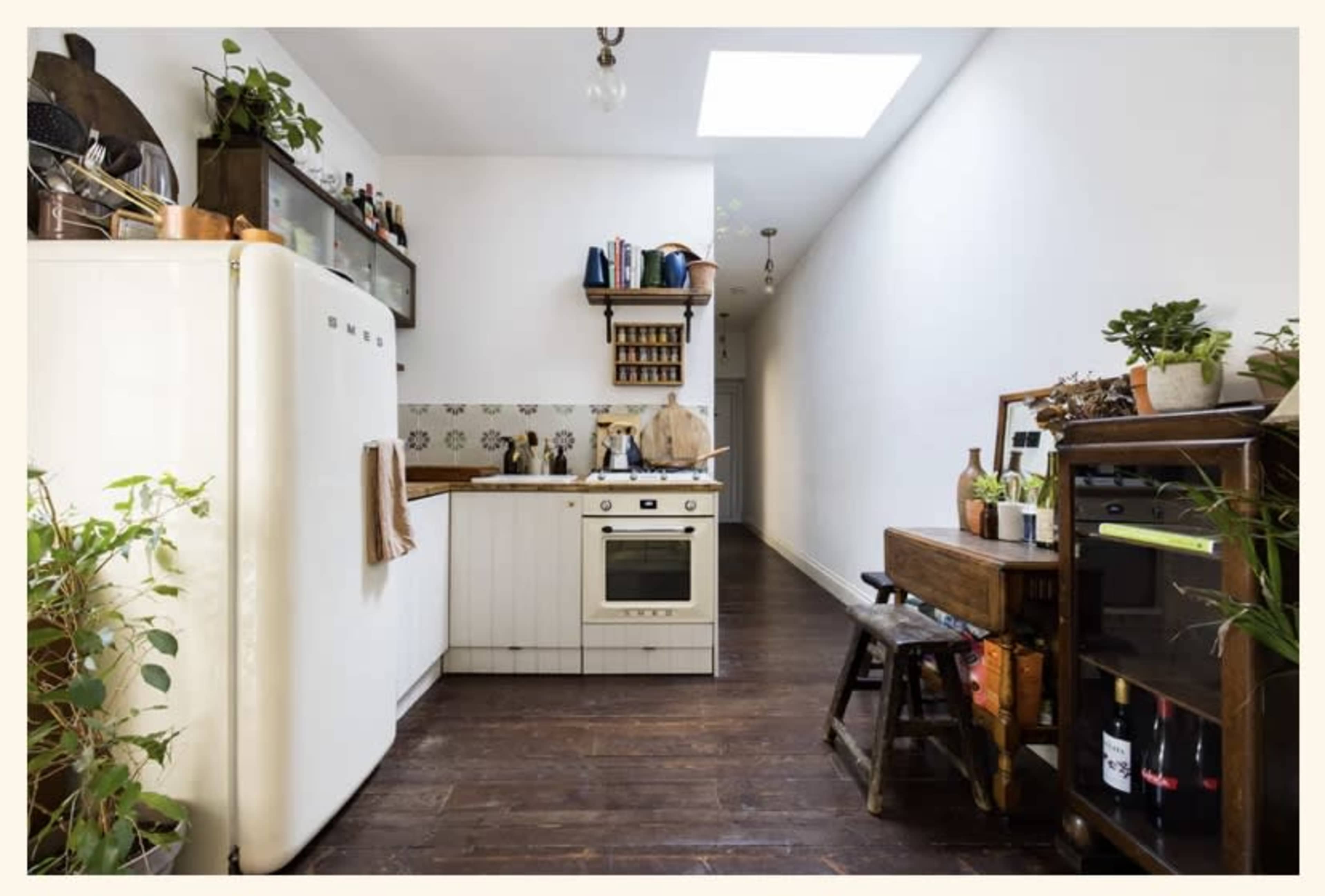 A narrow kitchen with a vintage white refrigerator, a stove, wooden shelves filled with kitchen items, and plants along the walls, creating an organized space.