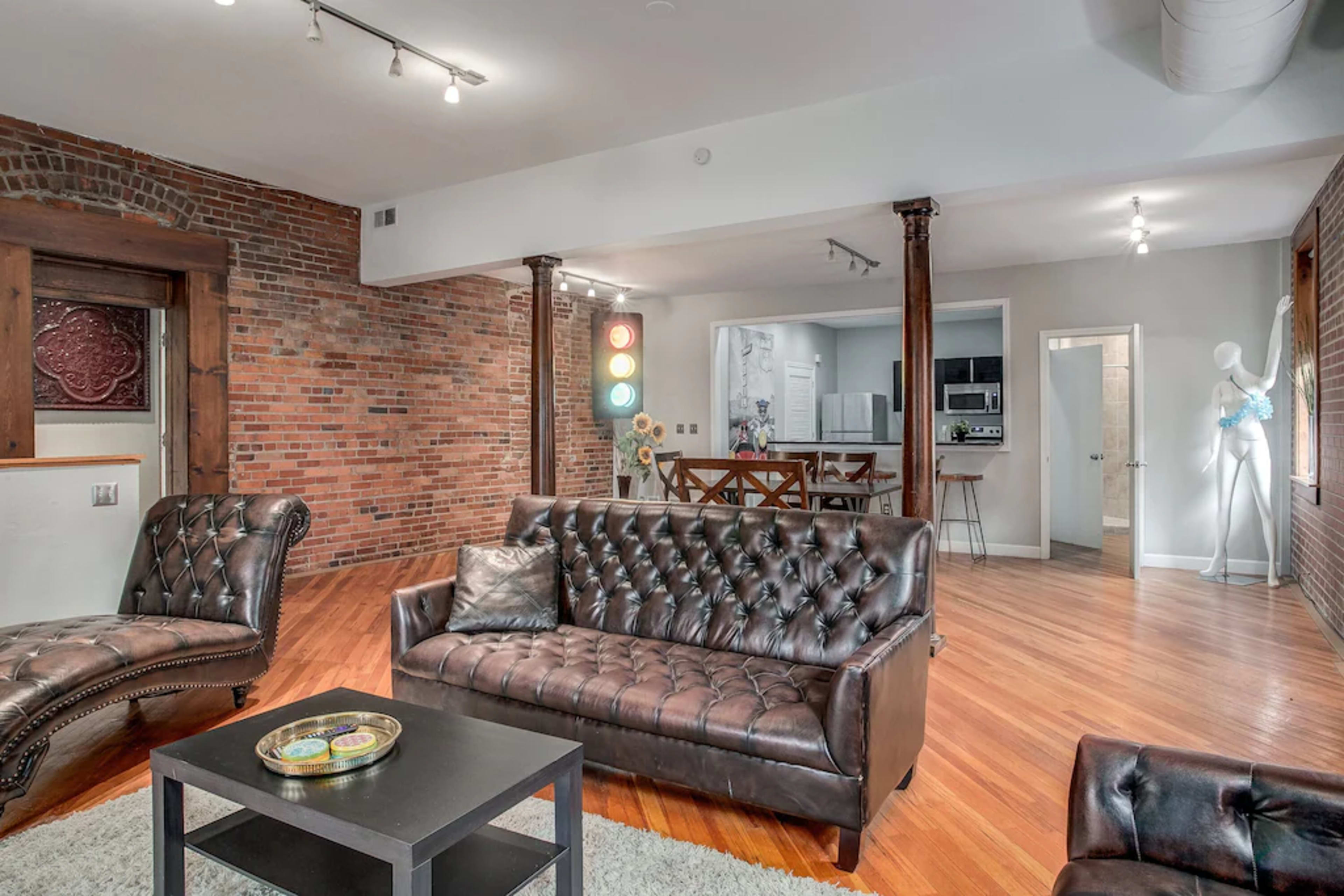 A spacious living area featuring a brown leather couch, a matching chair, hardwood flooring, exposed brick walls, and a glimpse of a kitchen in the background.