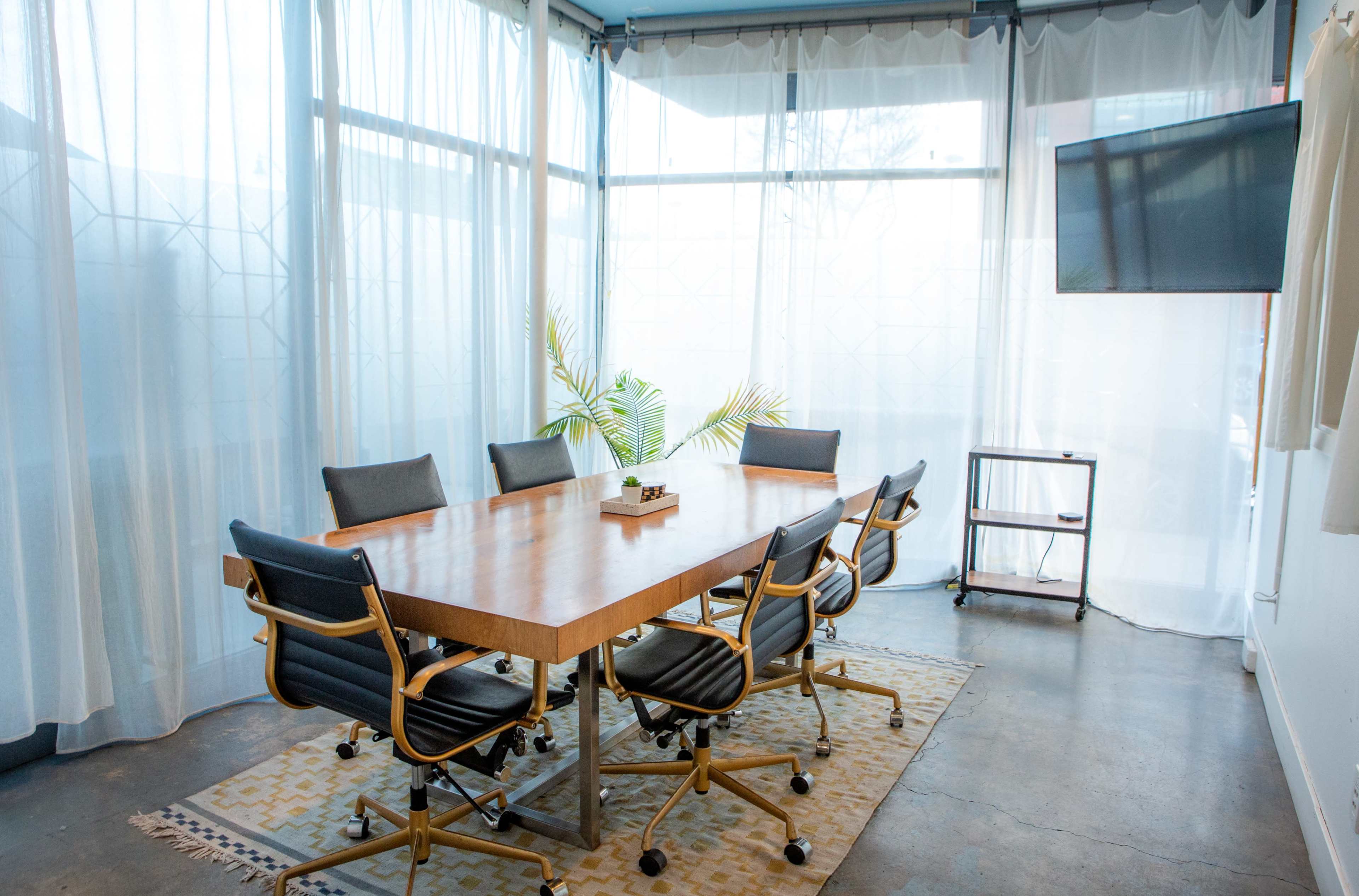 A modern conference room features a wooden table surrounded by black rolling chairs, with large windows adorned with sheer curtains and a small plant in the center.