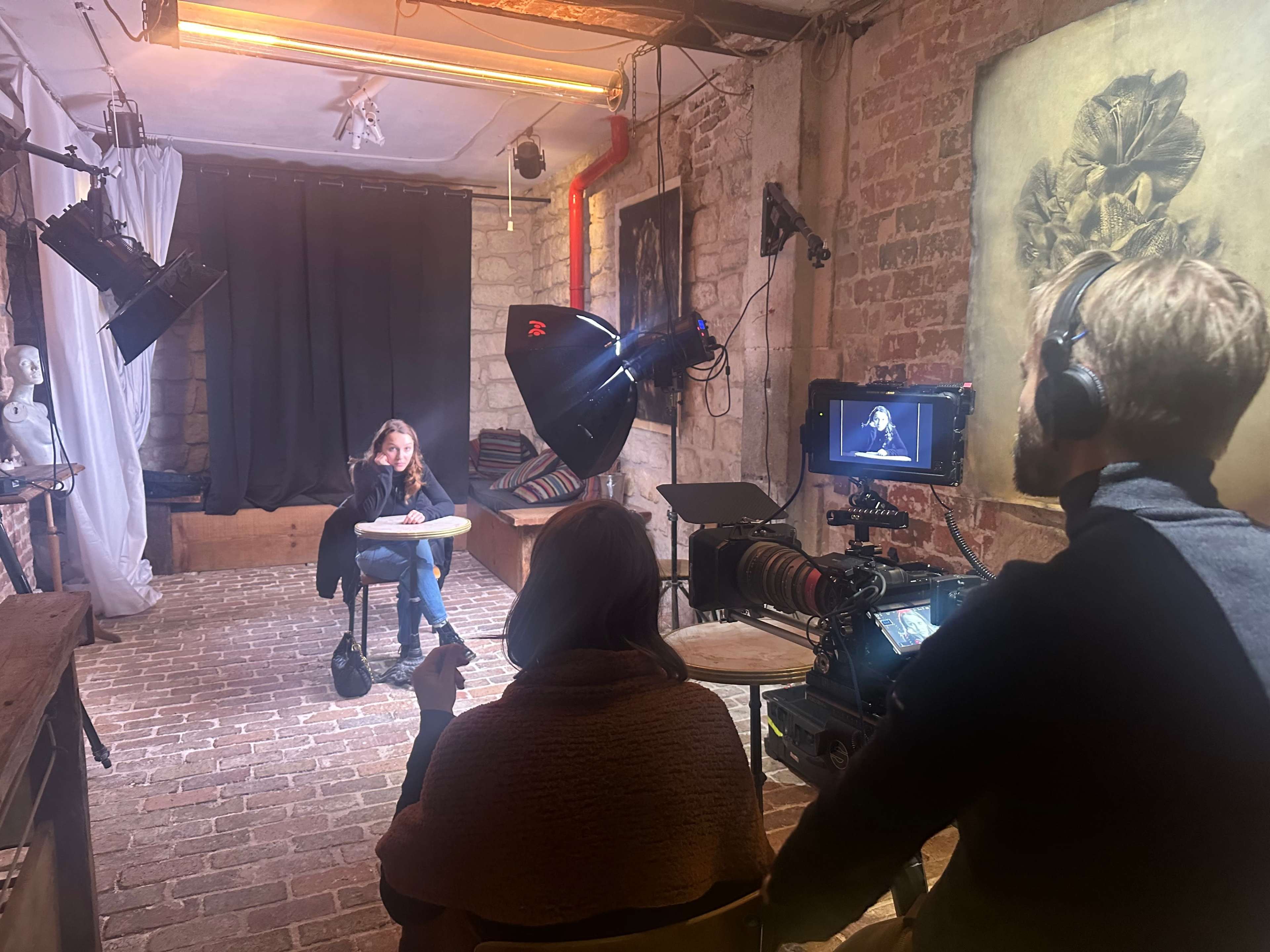 A woman sits at a table in a studio setting while a camera crew prepares to film her against a backdrop of black curtains.