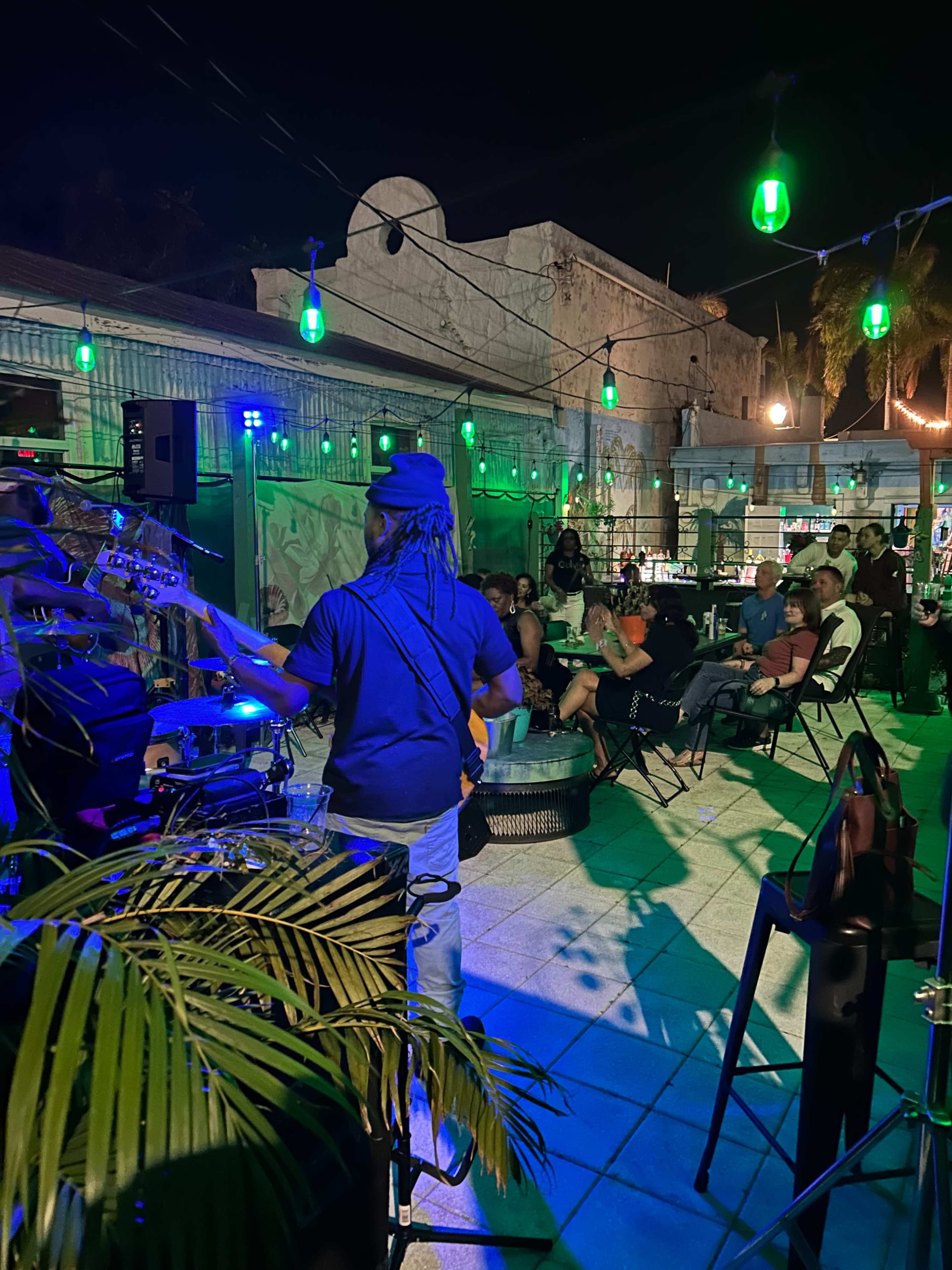 A musician plays guitar on stage while an audience enjoys the performance at an outdoor venue illuminated by colorful lights.