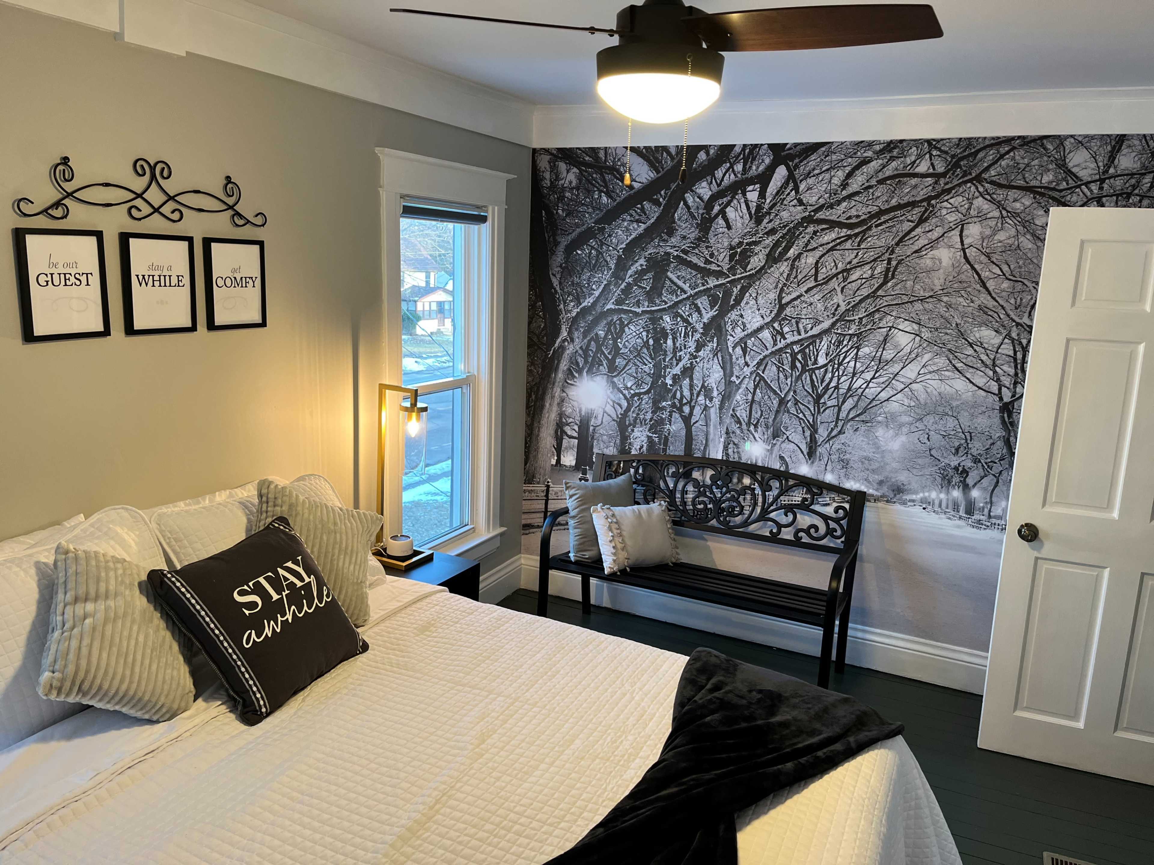 A cozy bedroom with a bed, a decorative bench, and a large black-and-white mural of a tree-lined street on one wall.