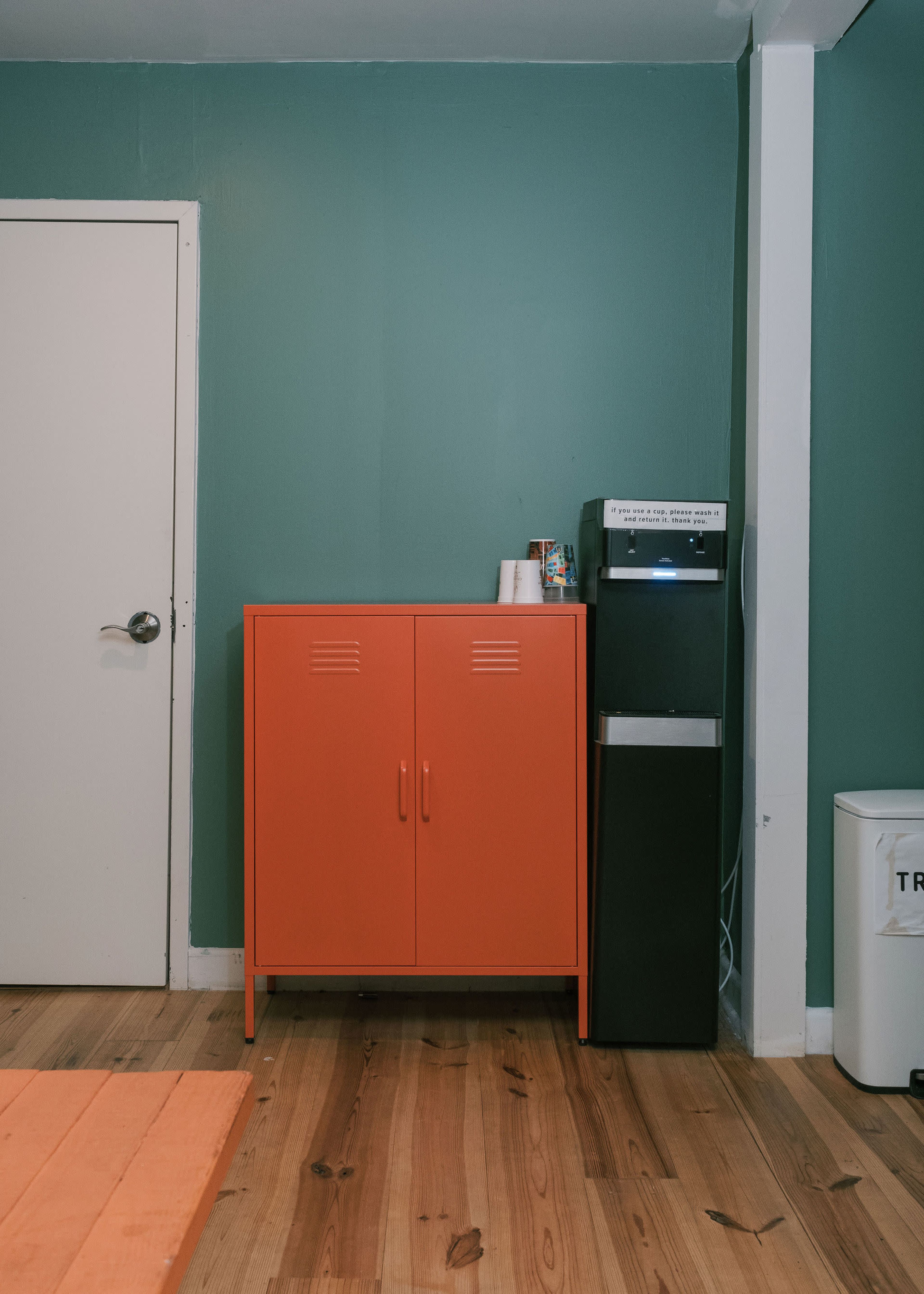 The image shows a room with a green wall, featuring an orange cabinet next to a black water cooler and a white trash can.