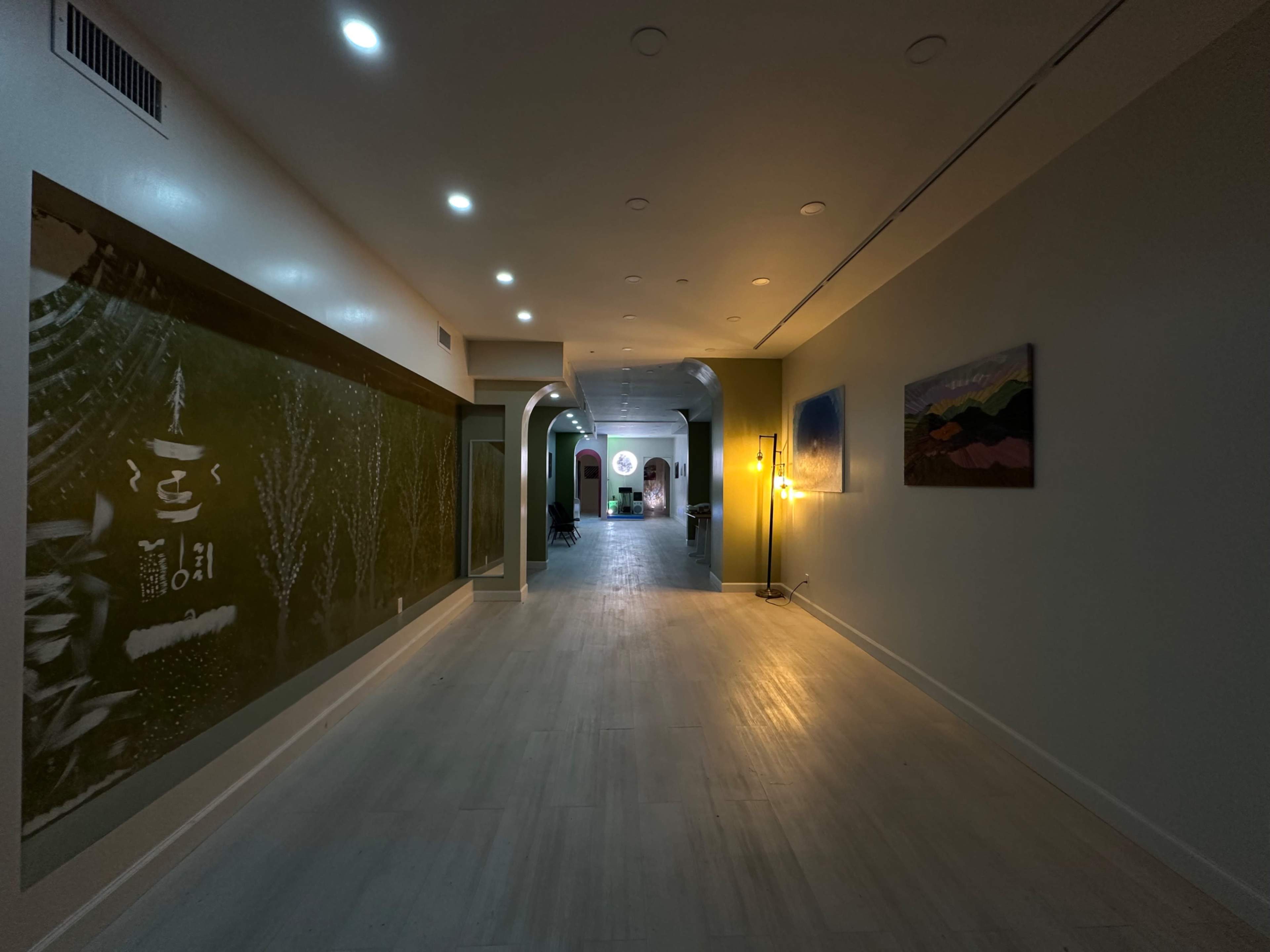 The image shows a long, well-lit hallway with a light-colored floor, featuring artwork on the walls and arched doorways leading to areas beyond.