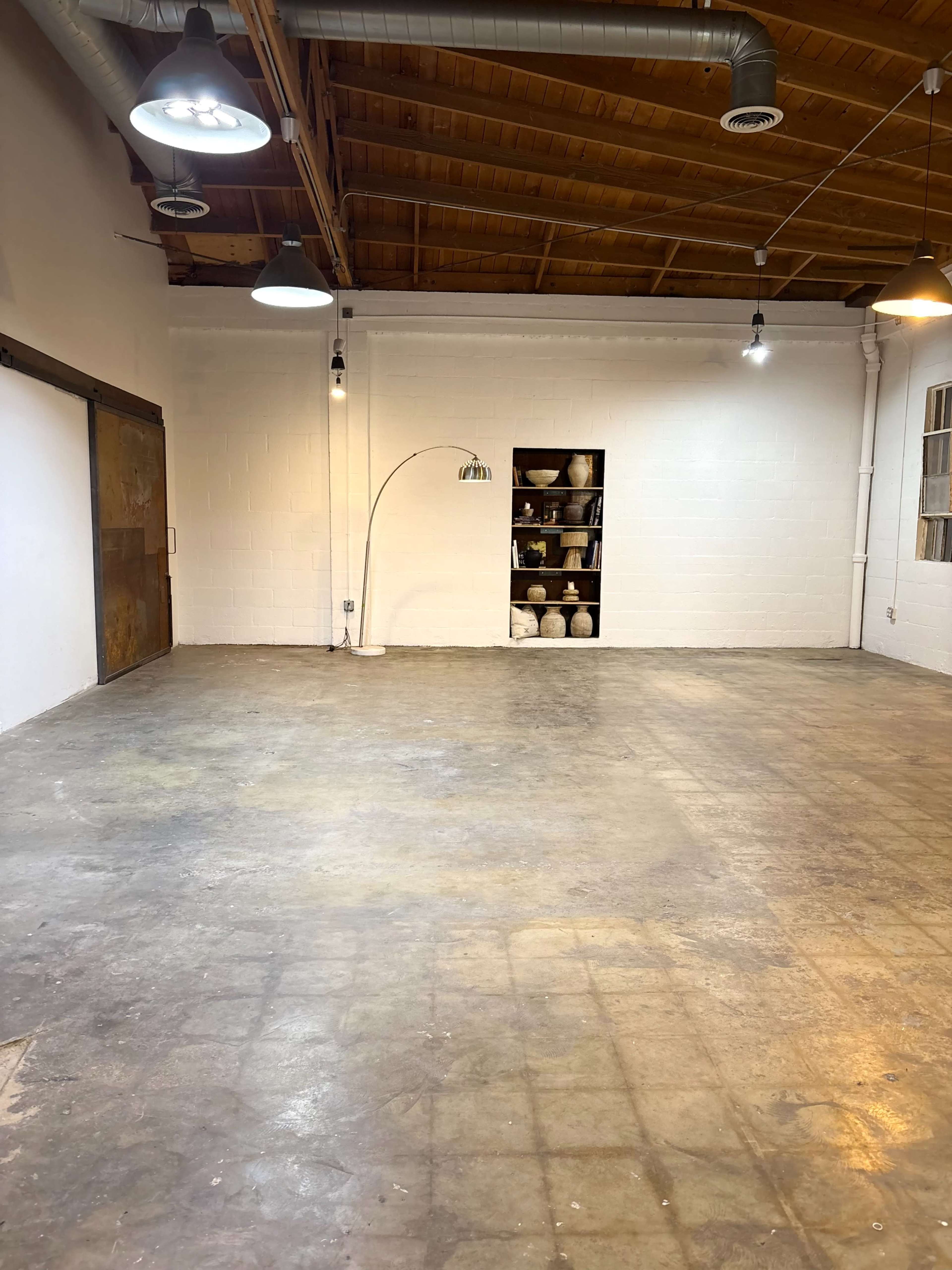 The image shows an empty industrial space with a concrete floor, wooden ceiling beams, and a wall-mounted shelf displaying ceramic items.