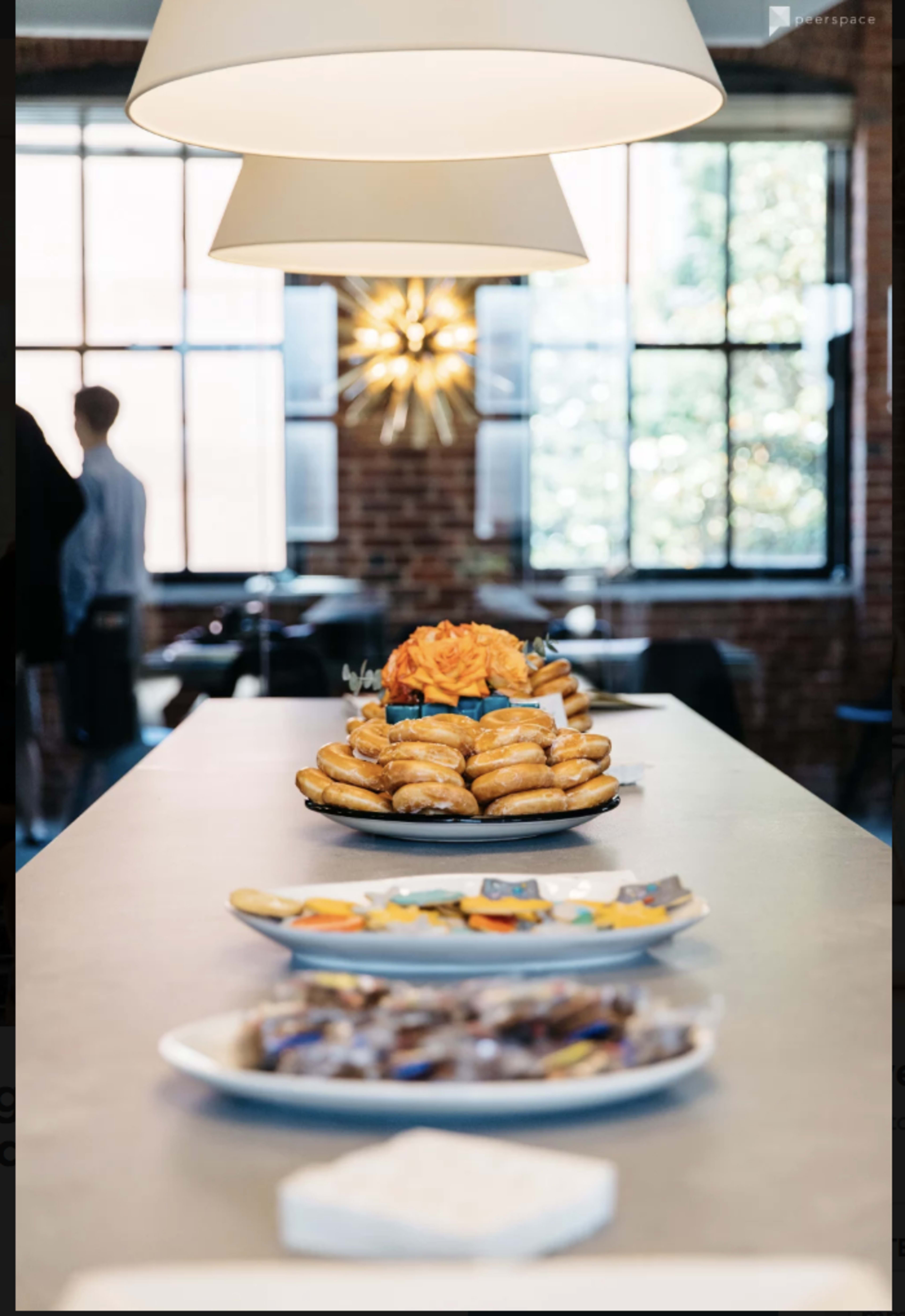 A long table features a platter of pastries at the center, flanked by smaller dishes of snacks, under hanging light fixtures in a brick wall setting.