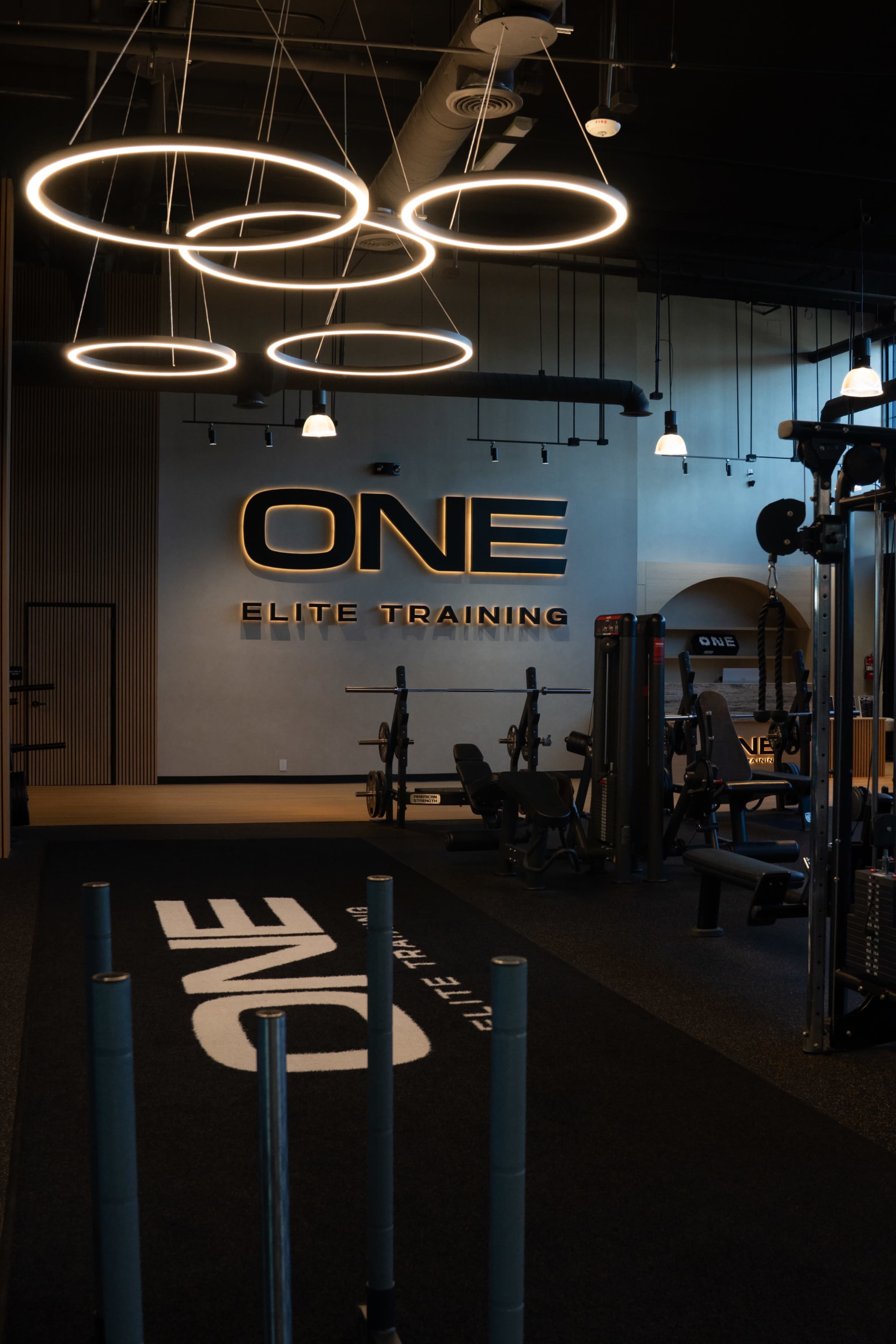 The image shows a modern gym interior featuring exercise equipment and a prominent "ONE ELITE TRAINING" sign on the wall, illuminated by circular light fixtures.
