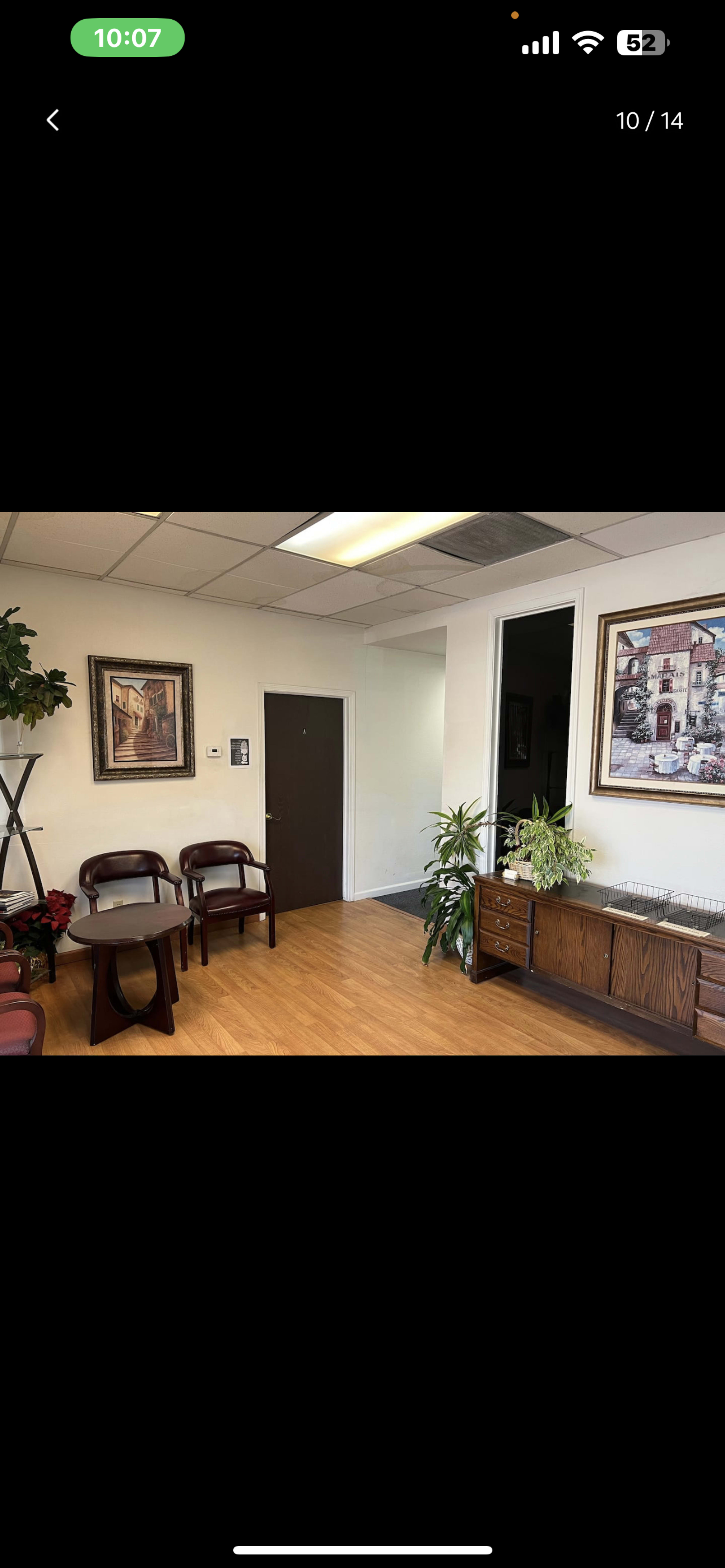 The image shows a reception area with two wooden chairs, a small table, and potted plants, alongside a painting on the wall and a cupboard.