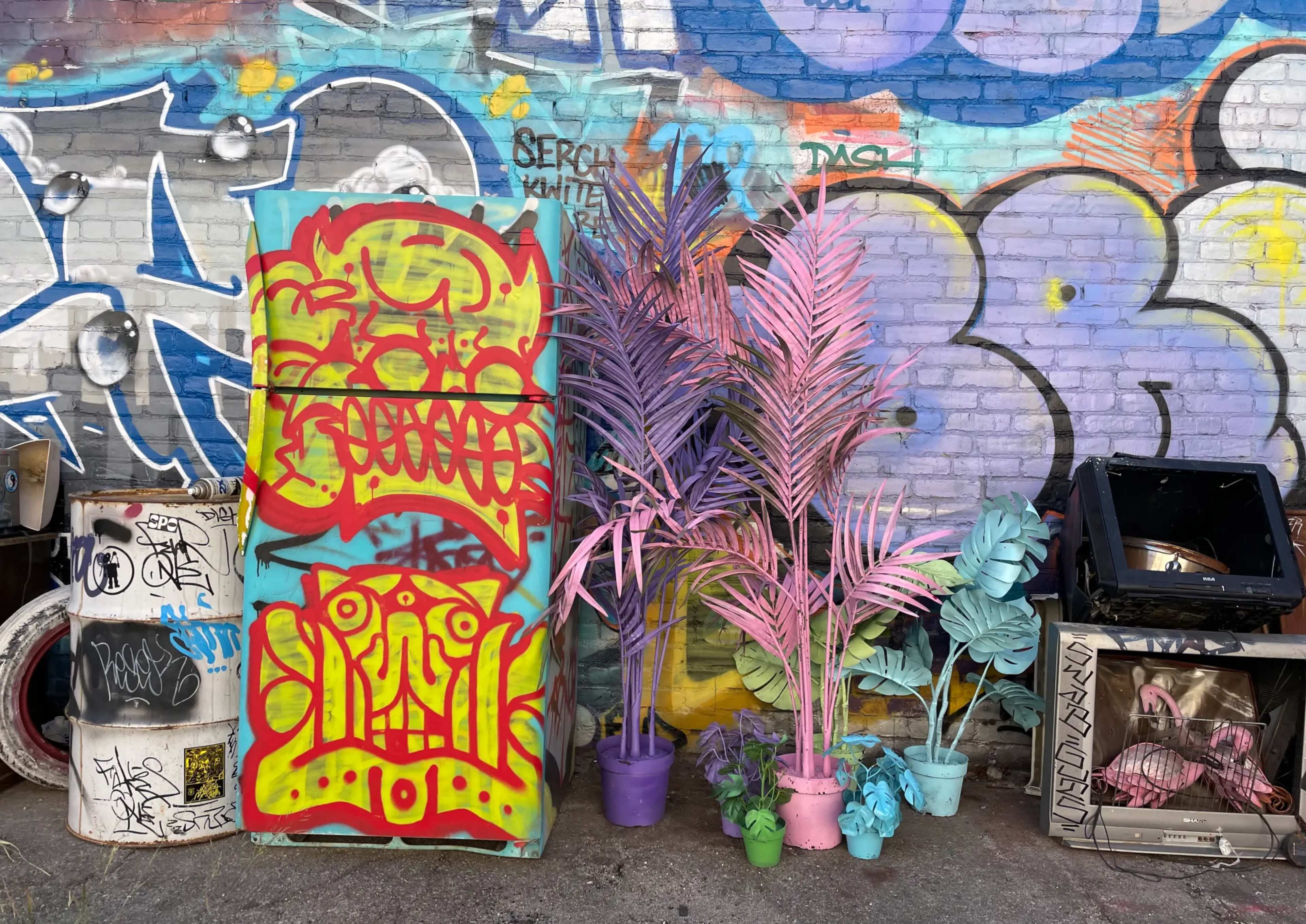The image shows a vibrant urban scene featuring a graffiti-covered wall, a colorful refrigerator adorned with graffiti, and potted plants in pastel colors placed beside it.