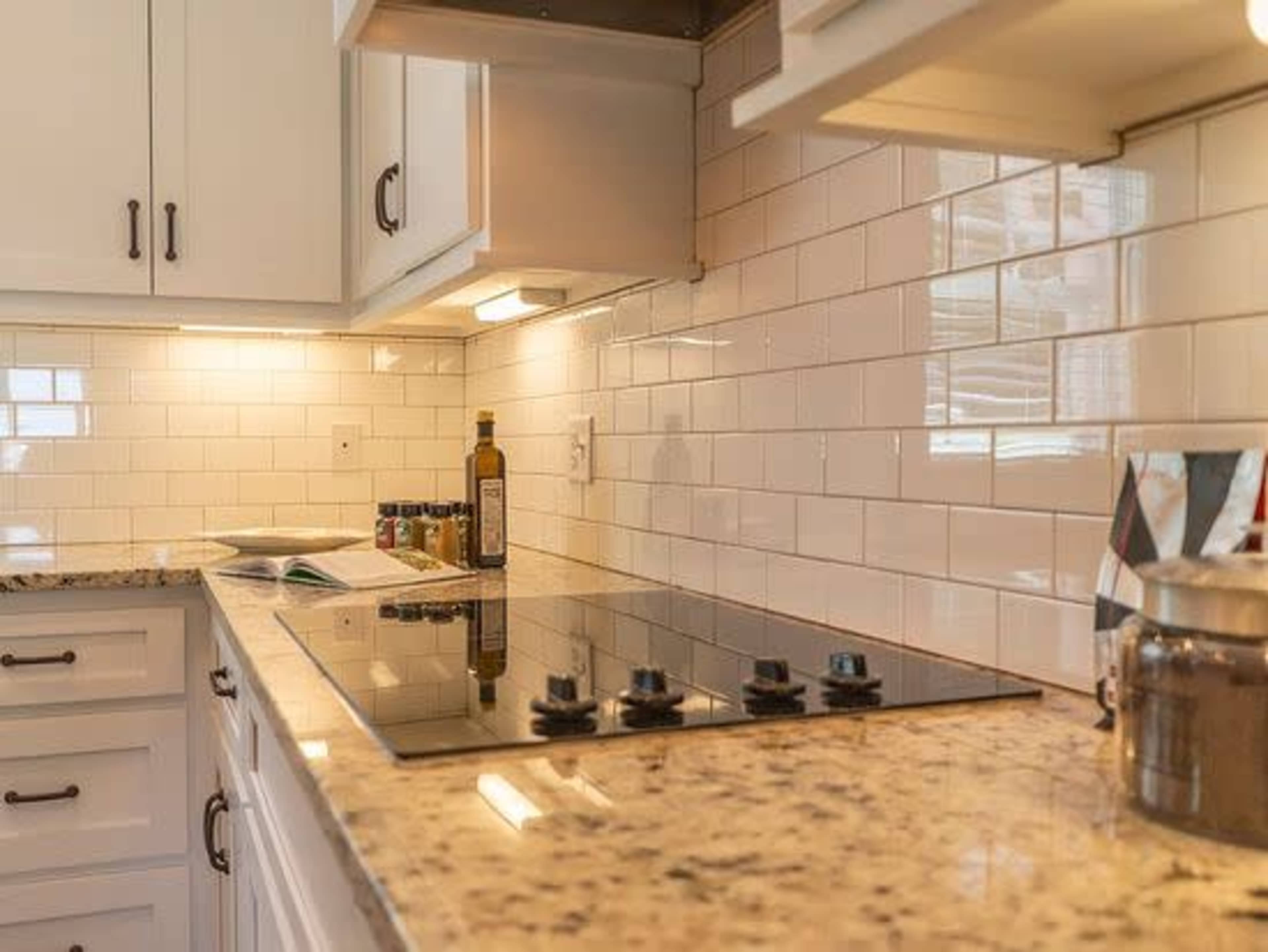 A modern kitchen countertop featuring a gas cooktop, neatly arranged spices, and a bottle of olive oil against a backdrop of white tile and light-colored cabinetry.