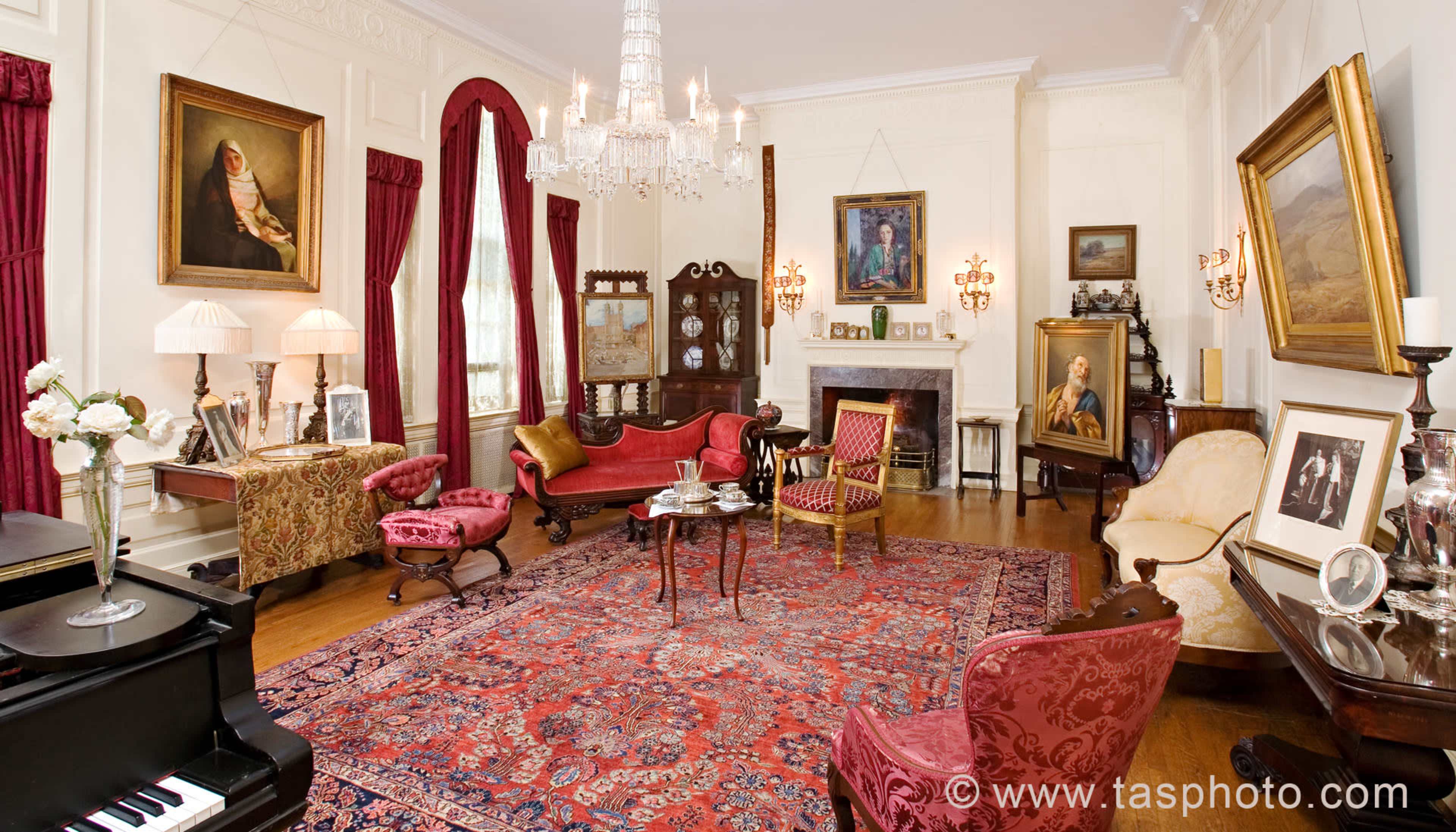 The scene shows a vintage living room with ornate furnishings, including a grand piano, multiple paintings on the walls, and a large, patterned rug.