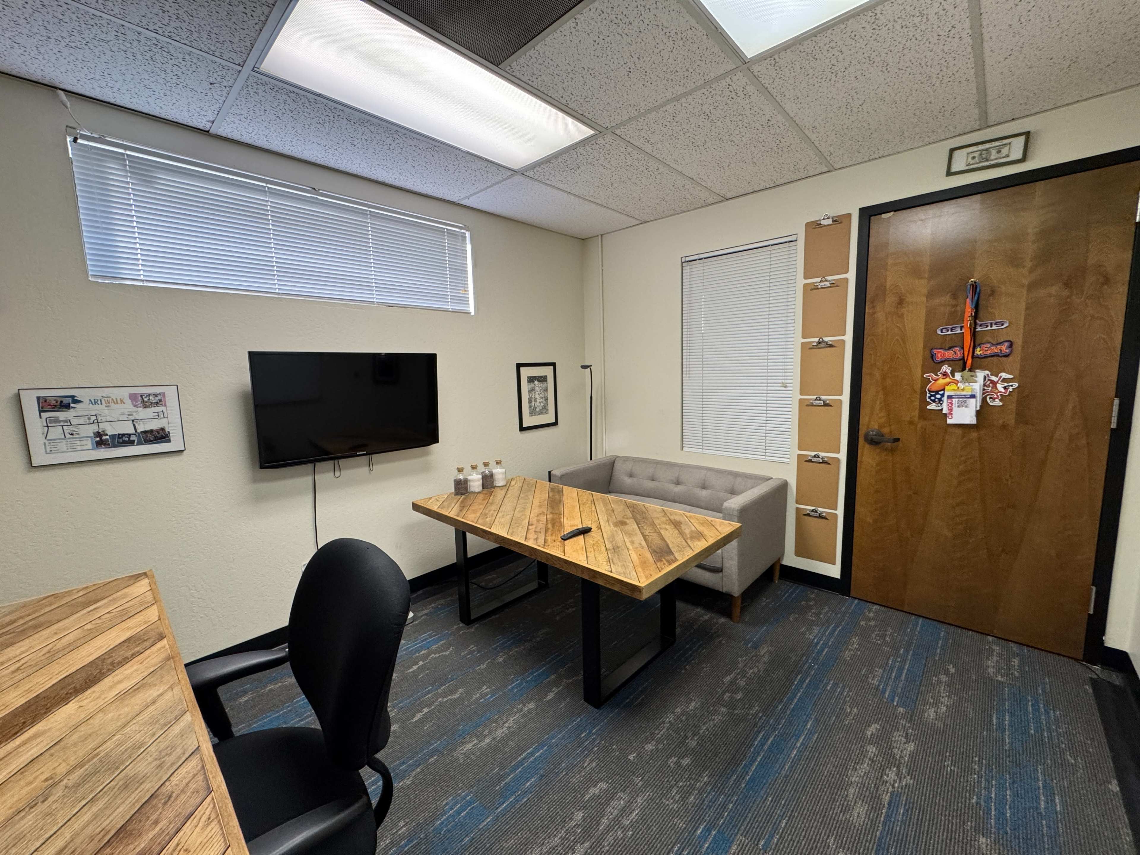 The image shows a small office space with a wooden table, a couch, a television mounted on the wall, and a door decorated with various items.