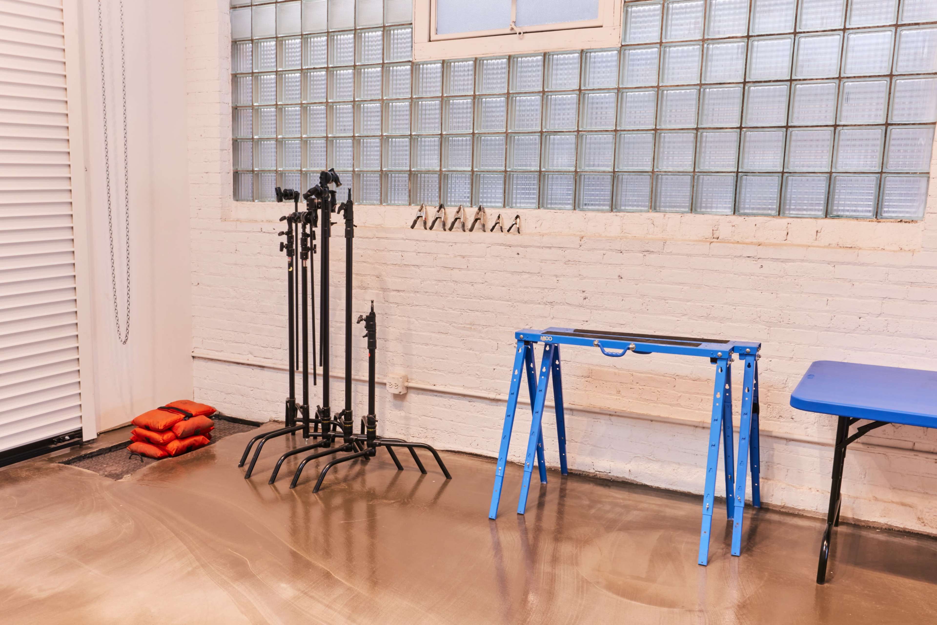 A workspace with a blue folding table, a set of black metal stands, and red sandbags on a polished floor, against a white brick wall with glass block windows.