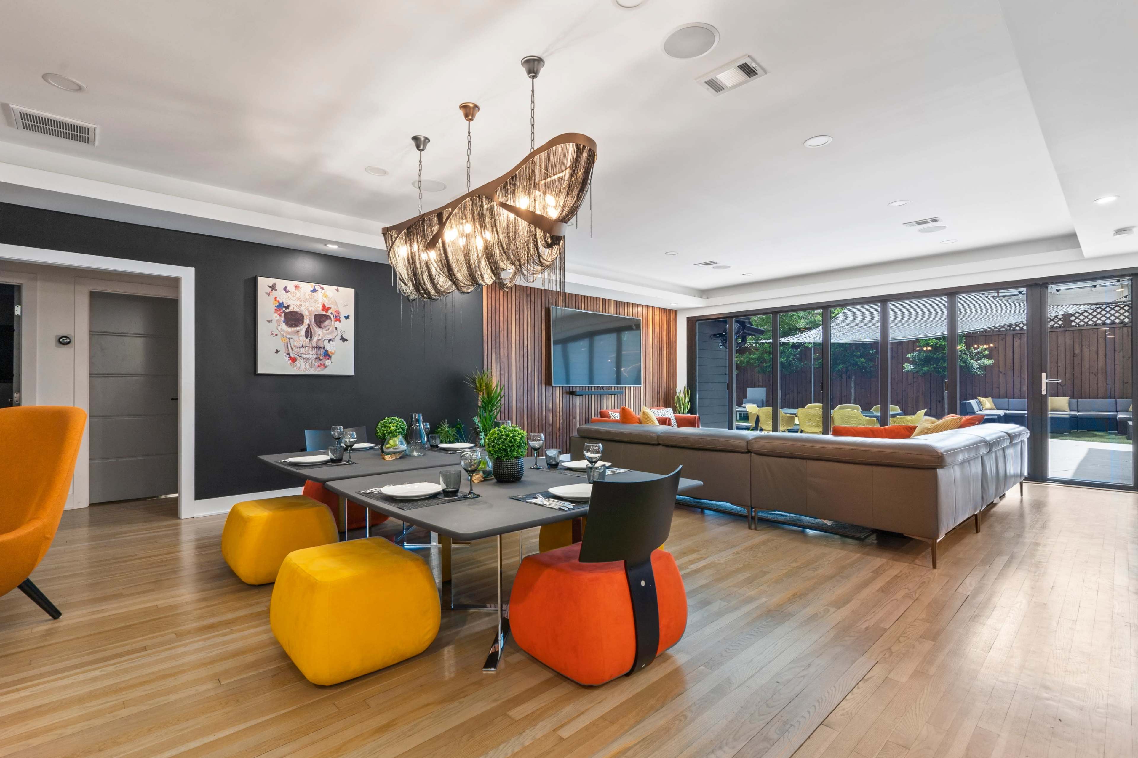 The image shows a modern living and dining area featuring a mix of colorful furniture, a large chandelier, and large glass doors leading to an outdoor space.