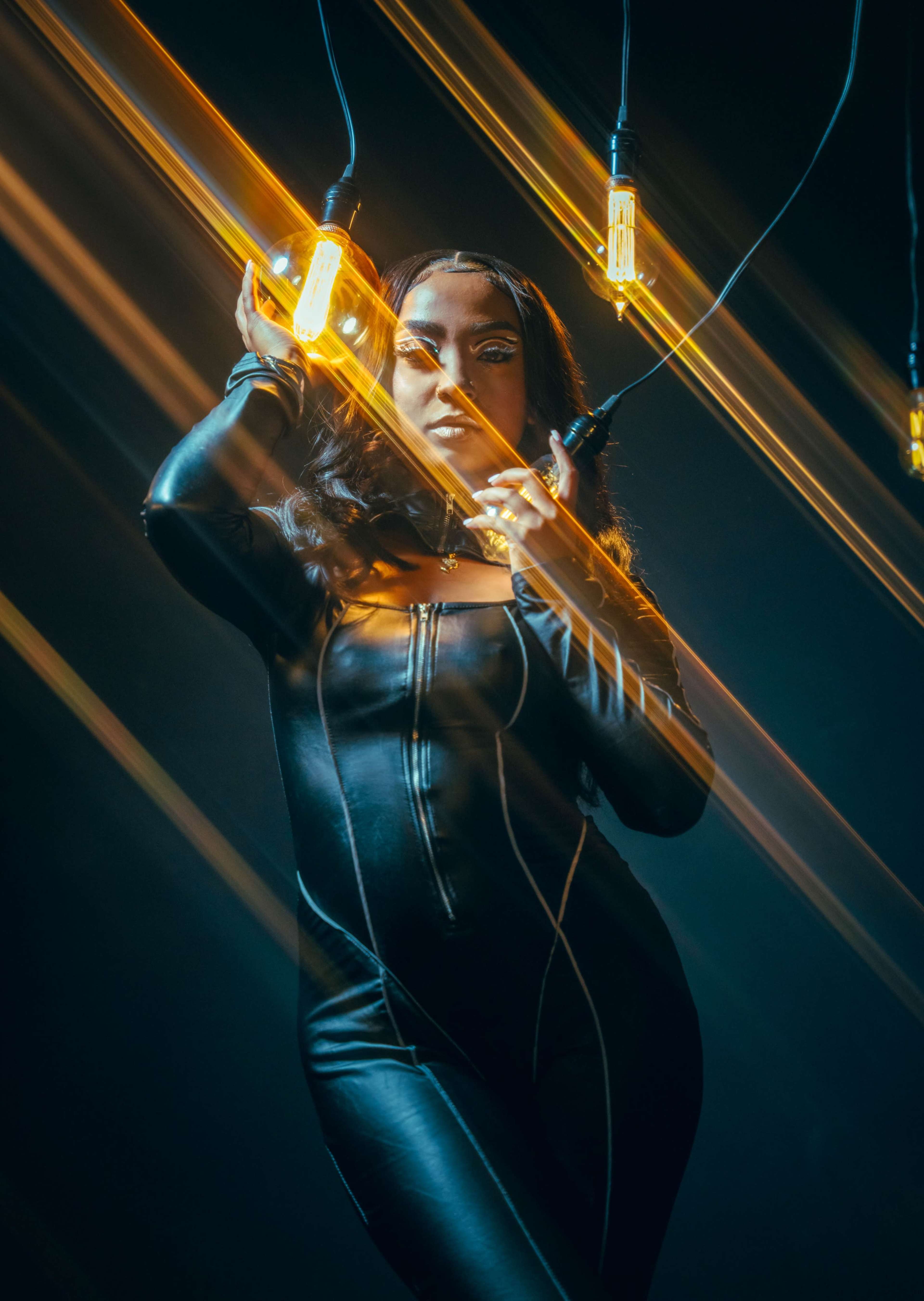 A person in a black outfit poses dramatically under hanging light bulbs against a dark background.