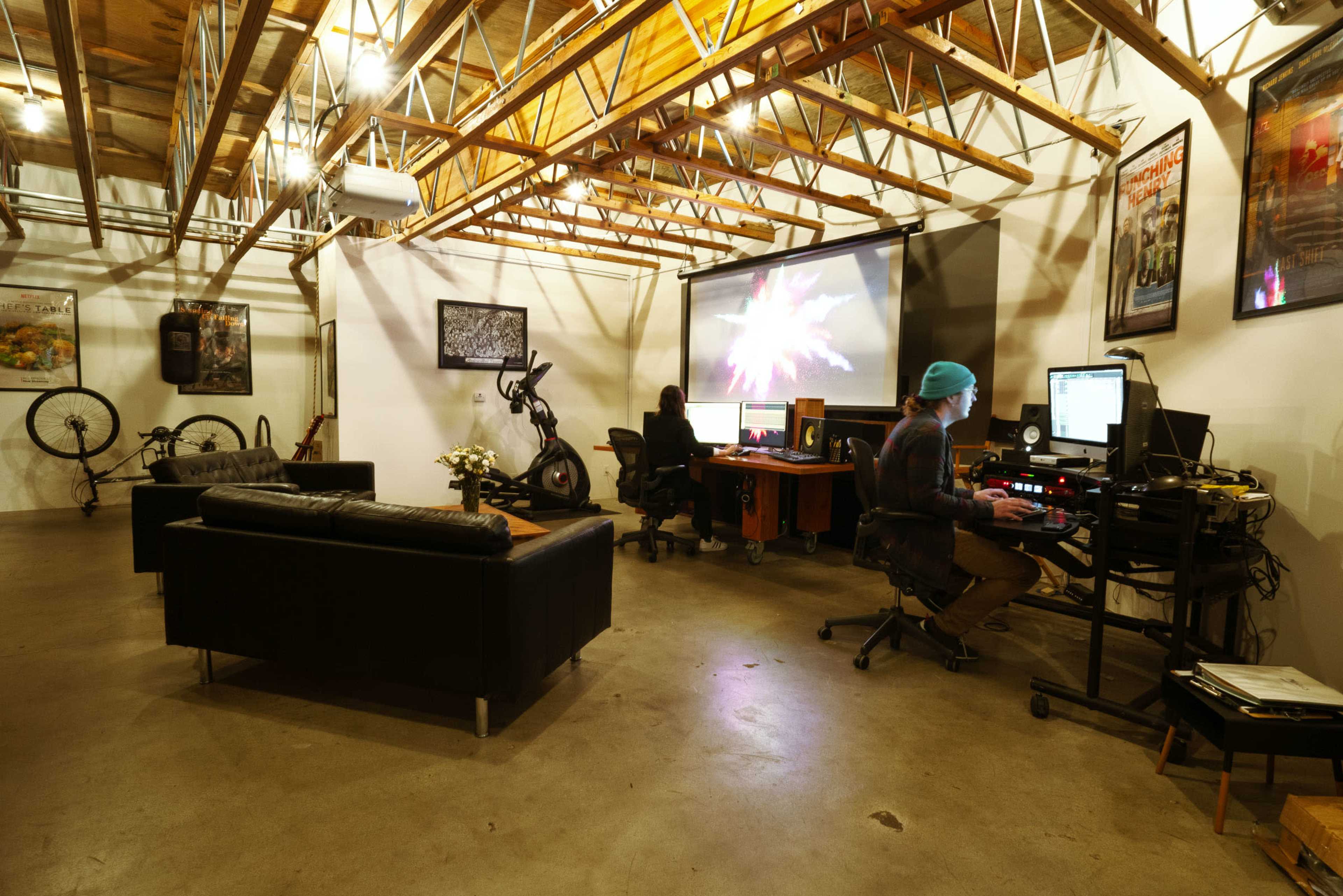 A modern office space with two workstations, a large screen displaying a graphic, and a seating area featuring black sofas.