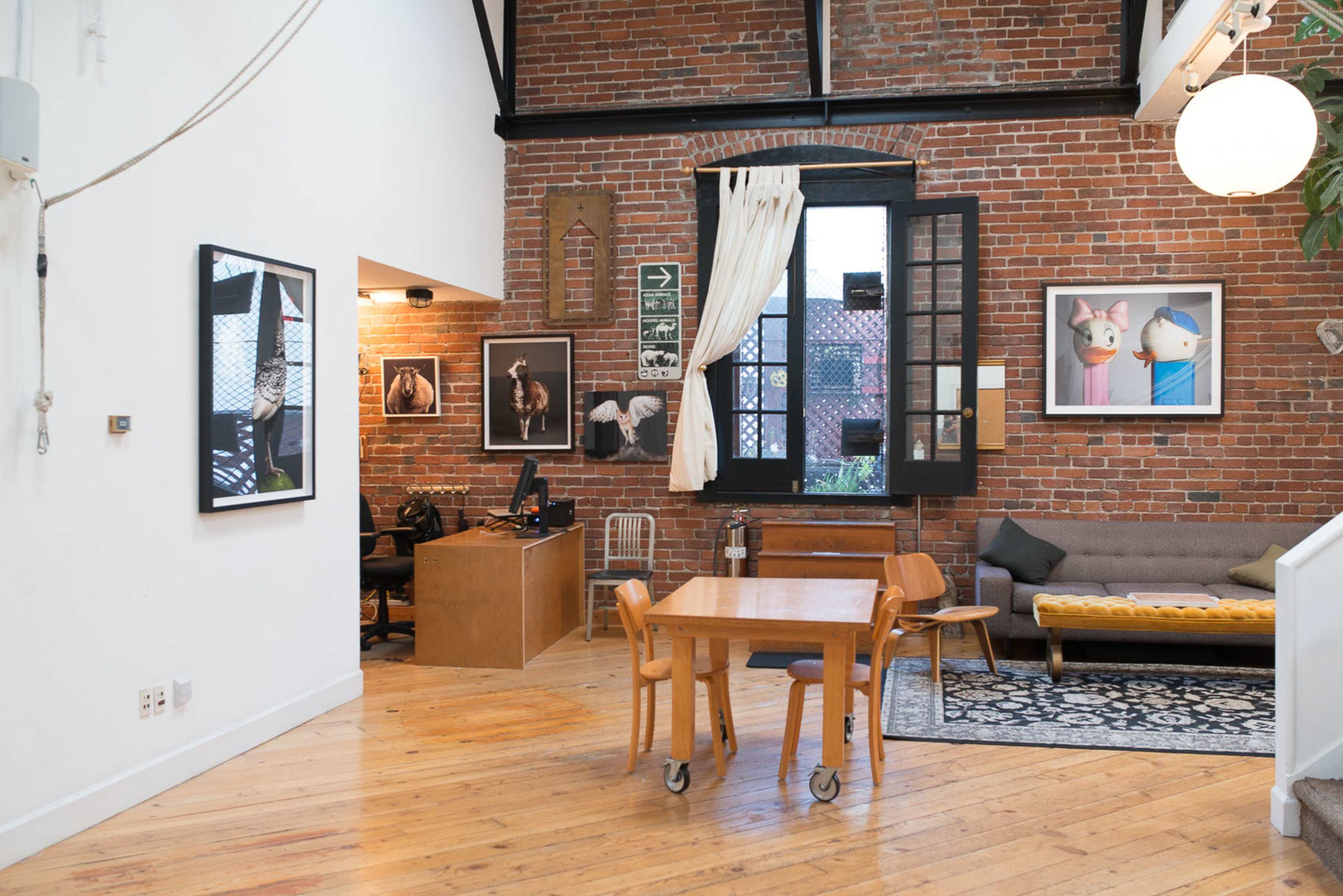 The image shows an interior of a spacious, contemporary room featuring exposed brick walls, a wooden table with chairs, and a workspace with a computer.