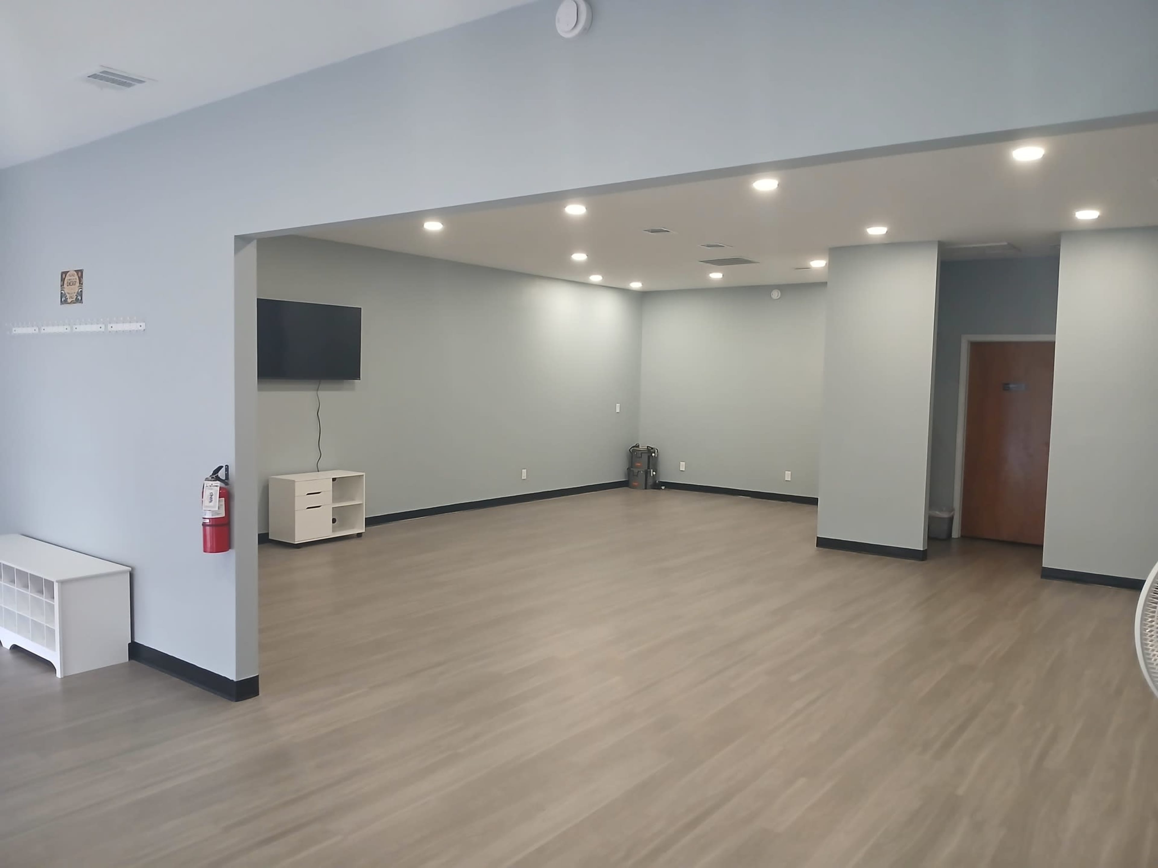 The image shows a spacious, empty room with light gray walls, wooden flooring, a television mounted on one wall, and minimal furnishings.