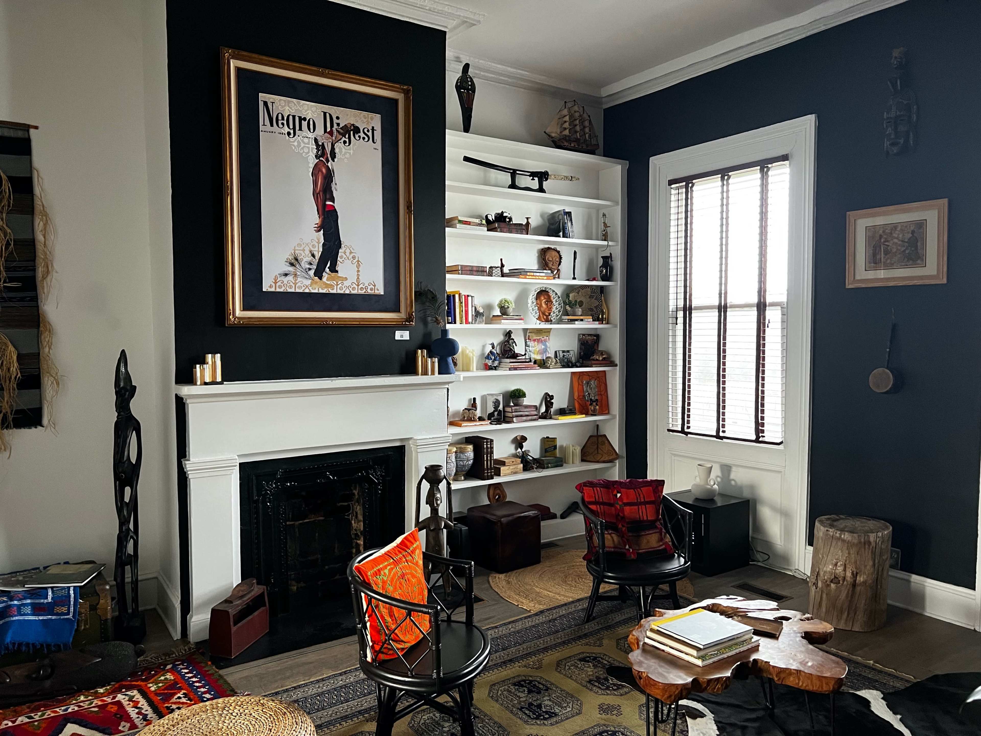 The room features a dark-colored wall with a prominent artwork, a white shelving unit filled with various decorations, a fireplace, and several chairs arranged around a textured rug.