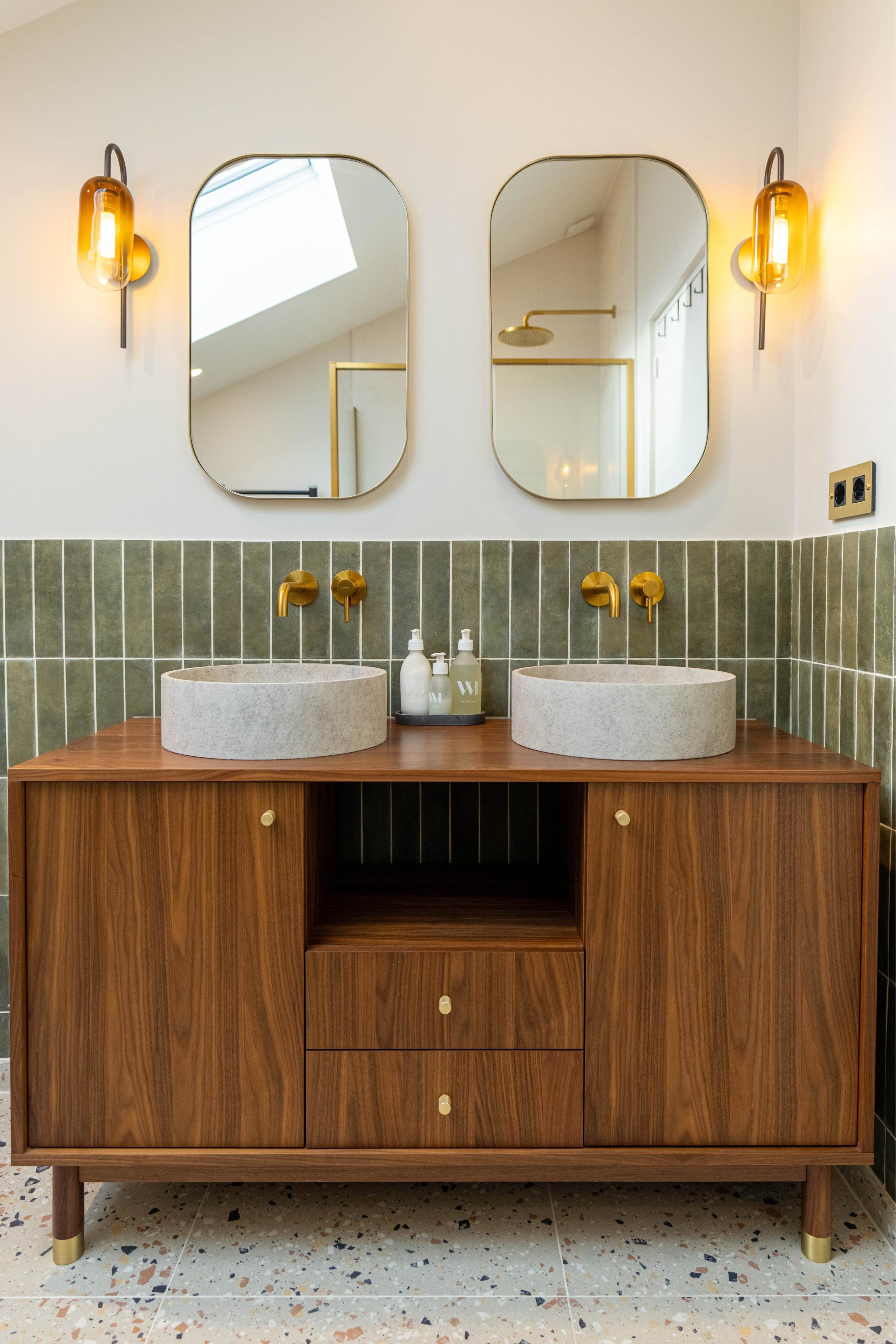 A modern bathroom features a wooden vanity with two circular stone sinks, set against green tiled walls and illuminated by wall-mounted light fixtures.