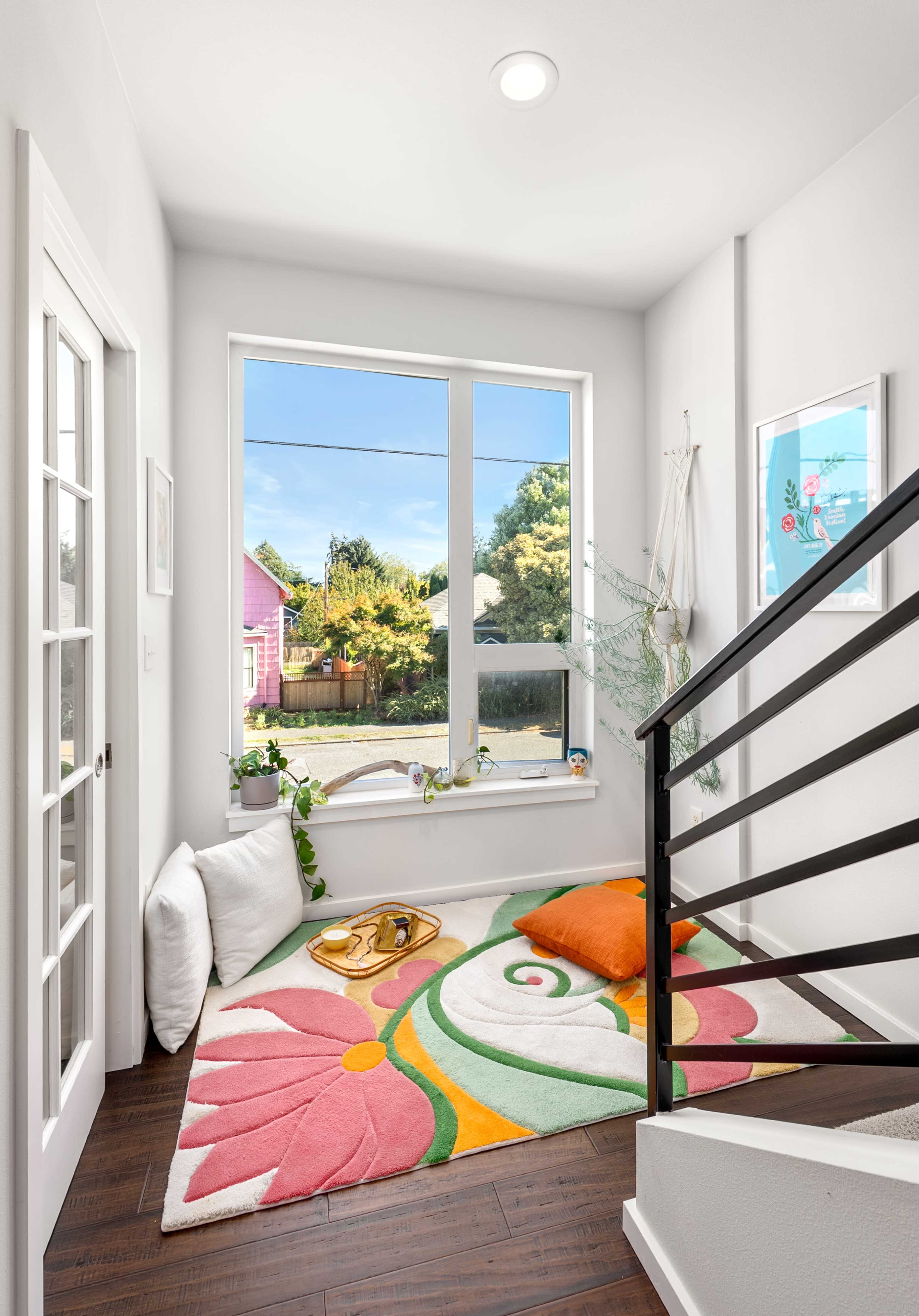 A colorful rug with floral designs is displayed on a hardwood floor in a well-lit entryway, featuring a large window overlooking a garden and a staircase with a modern railing.