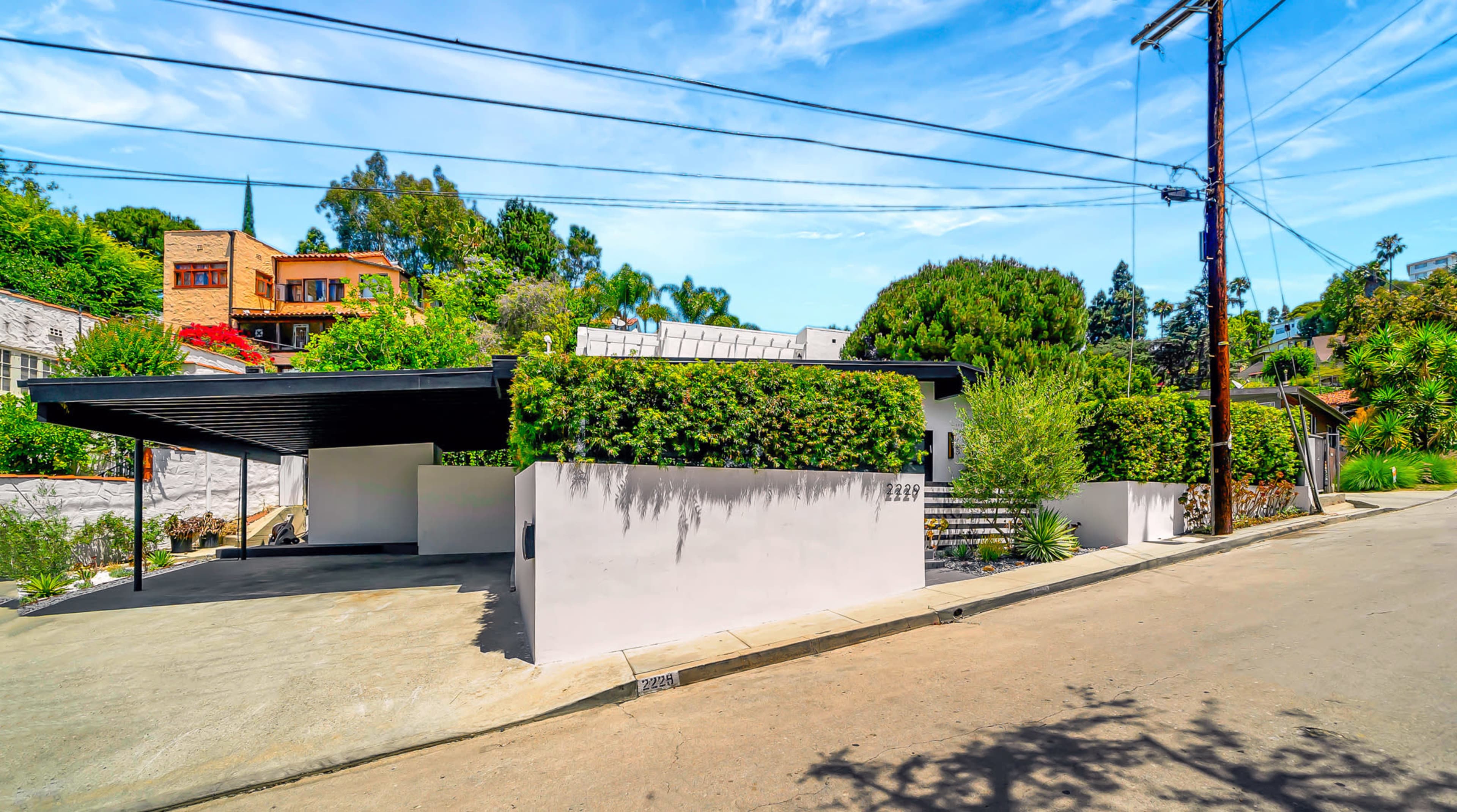 The image shows a modern house with a landscaped exterior located at the end of a sloped street, featuring a carport and greenery along the walls.