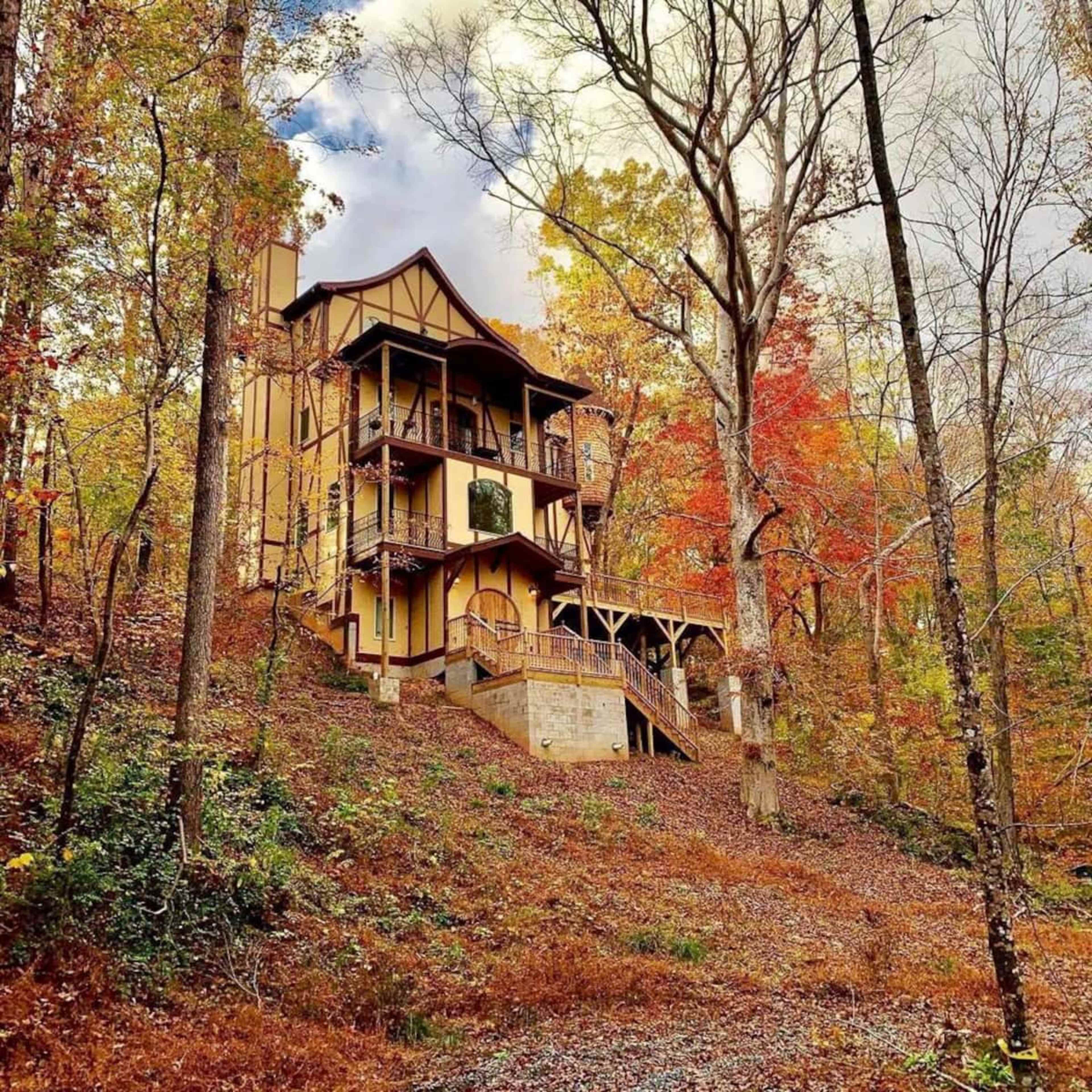 A multilevel house with a mix of wood and stone construction is situated on a hillside surrounded by trees displaying autumn foliage.