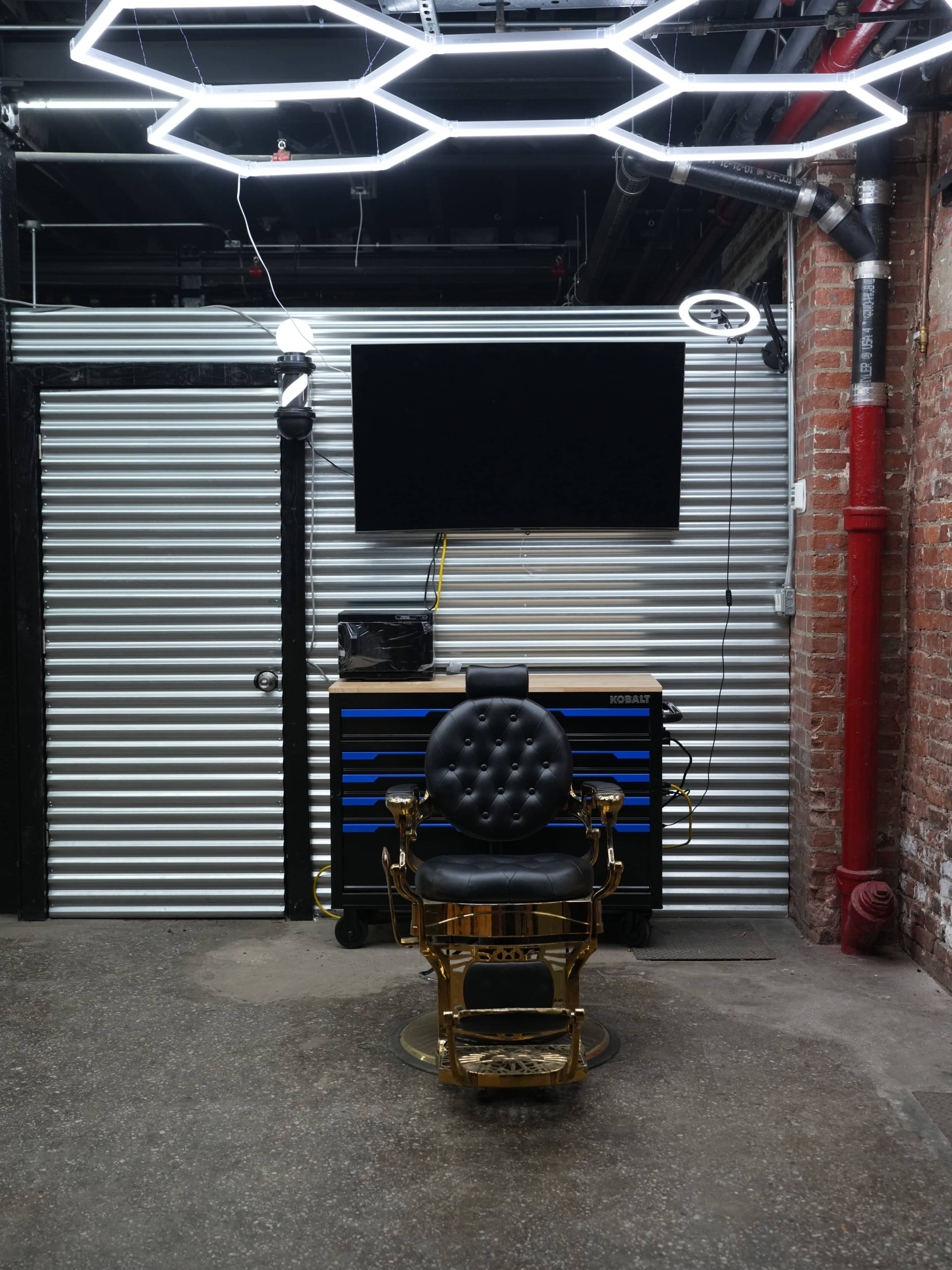 The image shows a barbershop setup with a vintage barber chair in front of a wall-mounted TV, surrounded by metal paneling and industrial-style lighting.