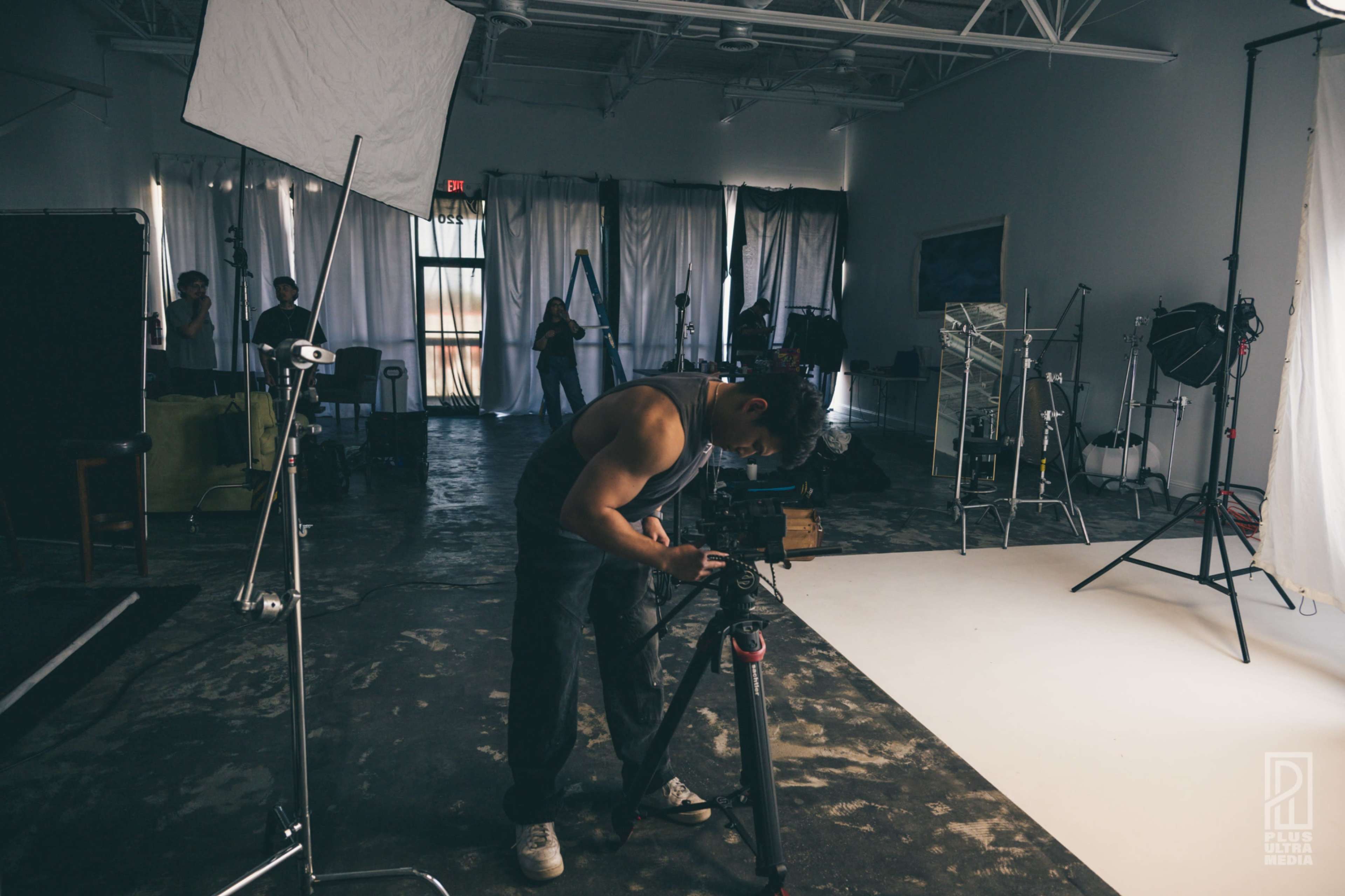 A photographer adjusts a camera on a tripod in a studio filled with lighting equipment and backdrops.