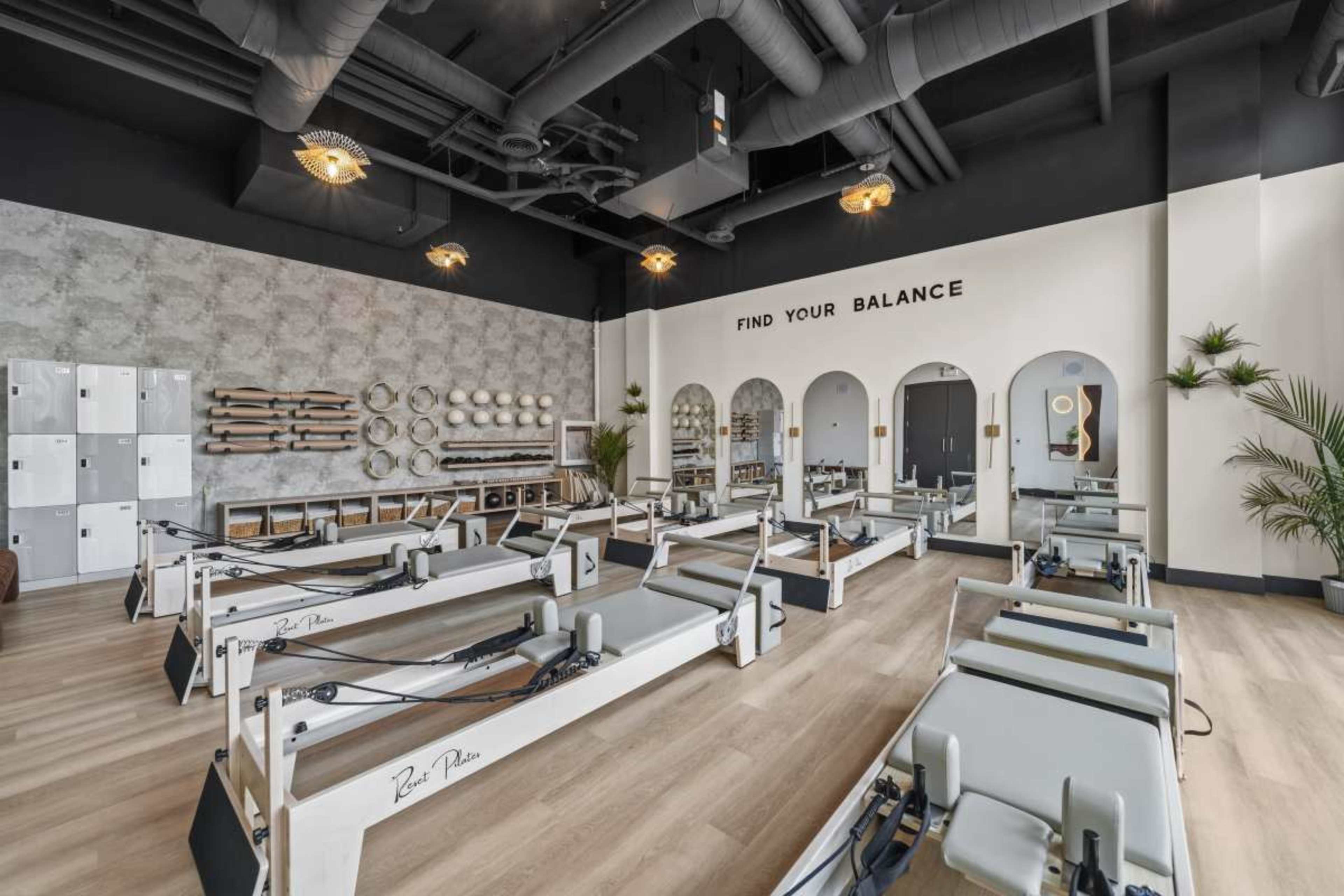 The image shows a modern fitness studio equipped with reformer Pilates machines, large windows, and minimalist decor.