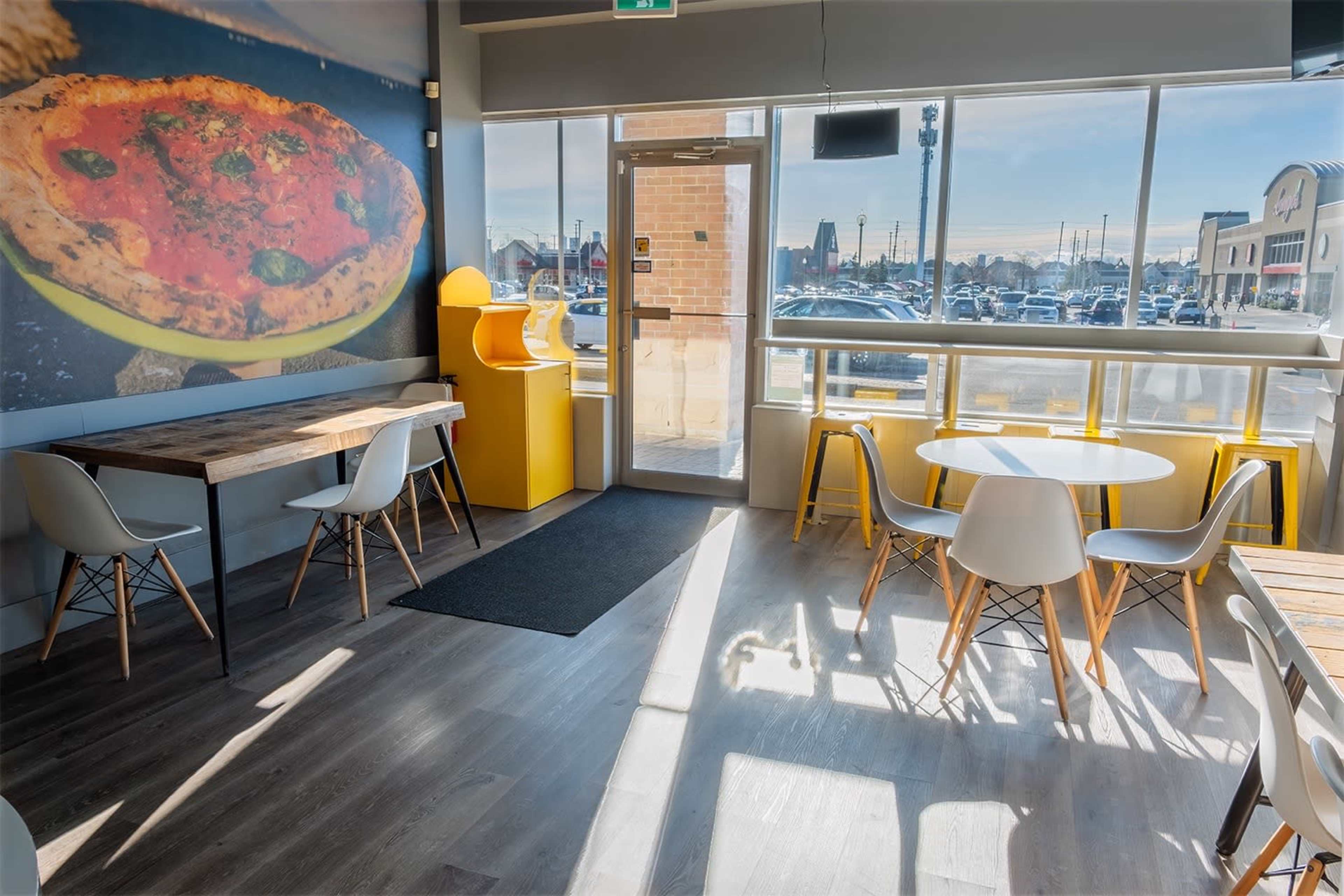 A modern restaurant interior with wooden tables, white chairs, and large windows that provide natural light, featuring a large pizza mural on one wall.