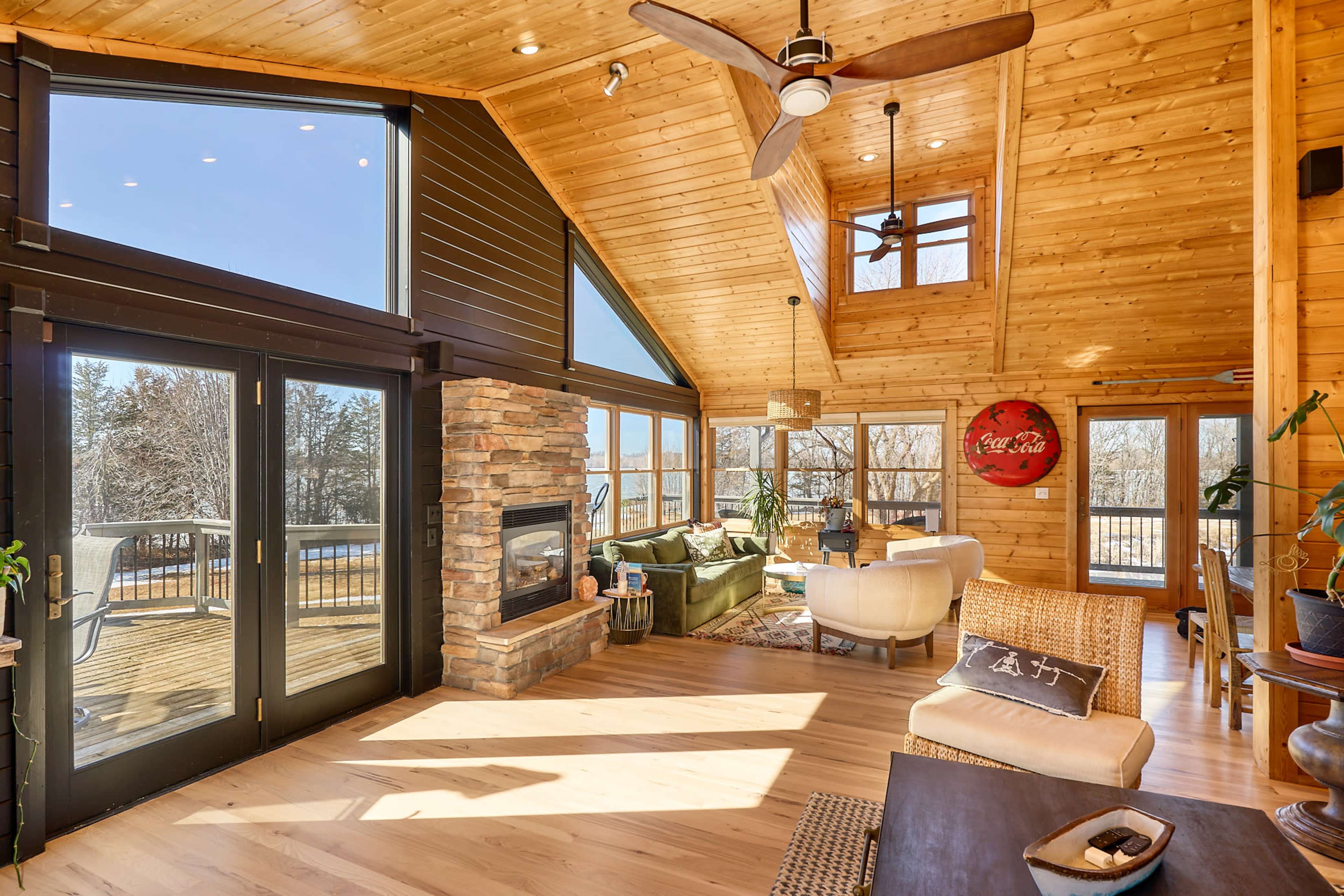 The image shows a spacious wooden living room with large windows, a stone fireplace, and modern furniture, looking out onto a scenic view.