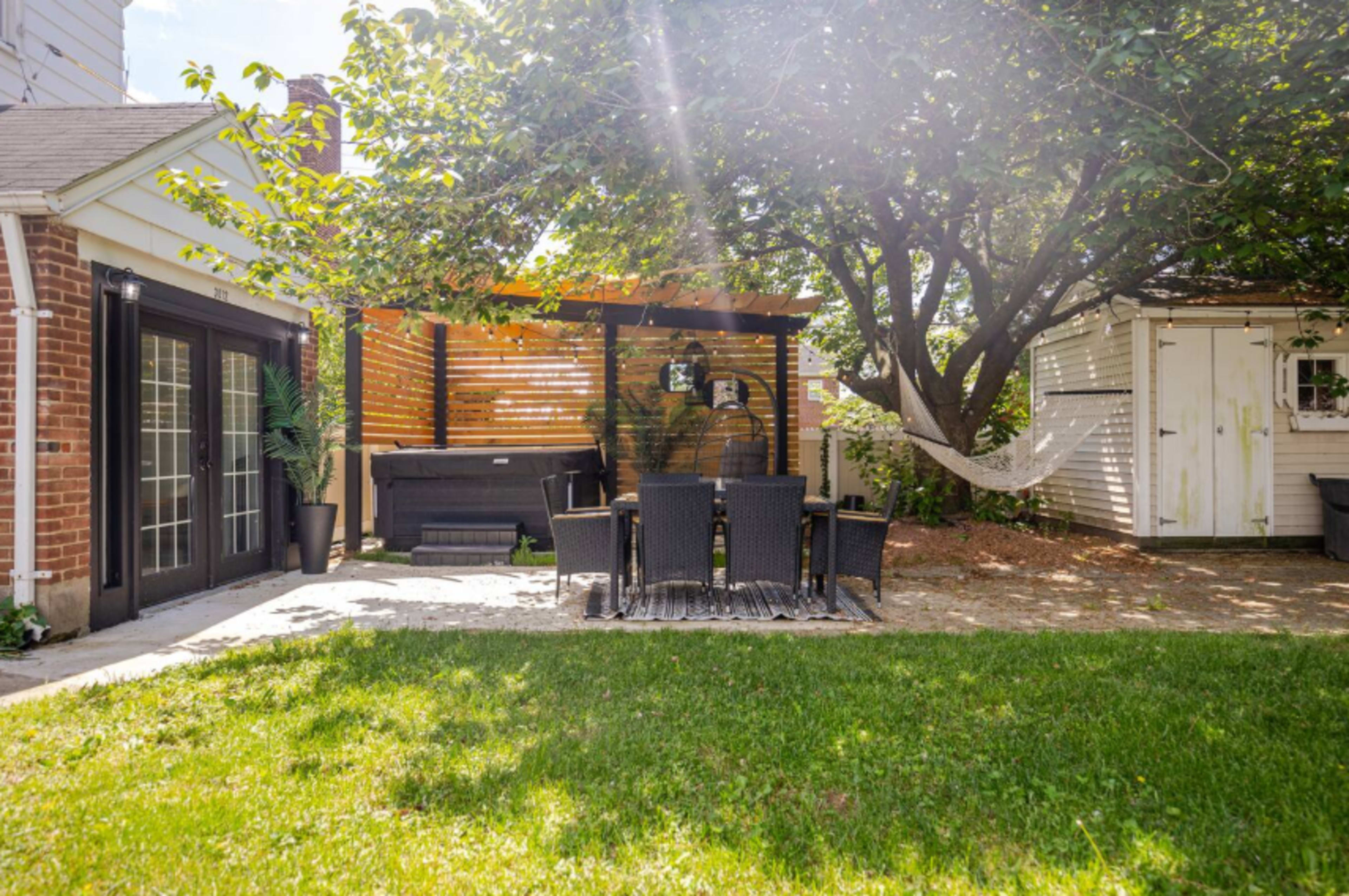 A shaded outdoor seating area features a black table and chairs under a wooden pergola beside a grassy yard and a white shed.