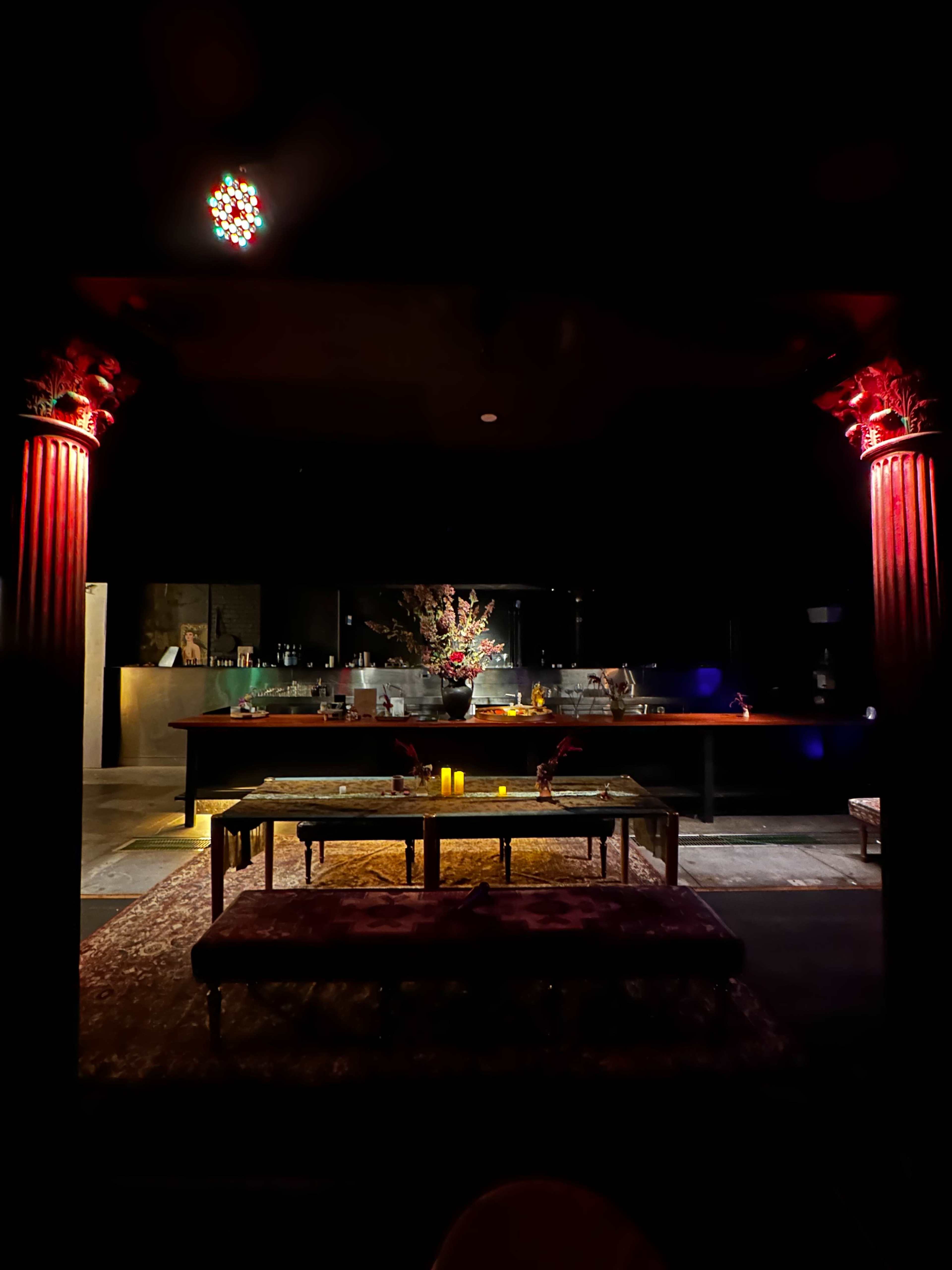 The image shows a dimly lit bar area with ornate pillars, a floral arrangement on the counter, and decorative seating in front.