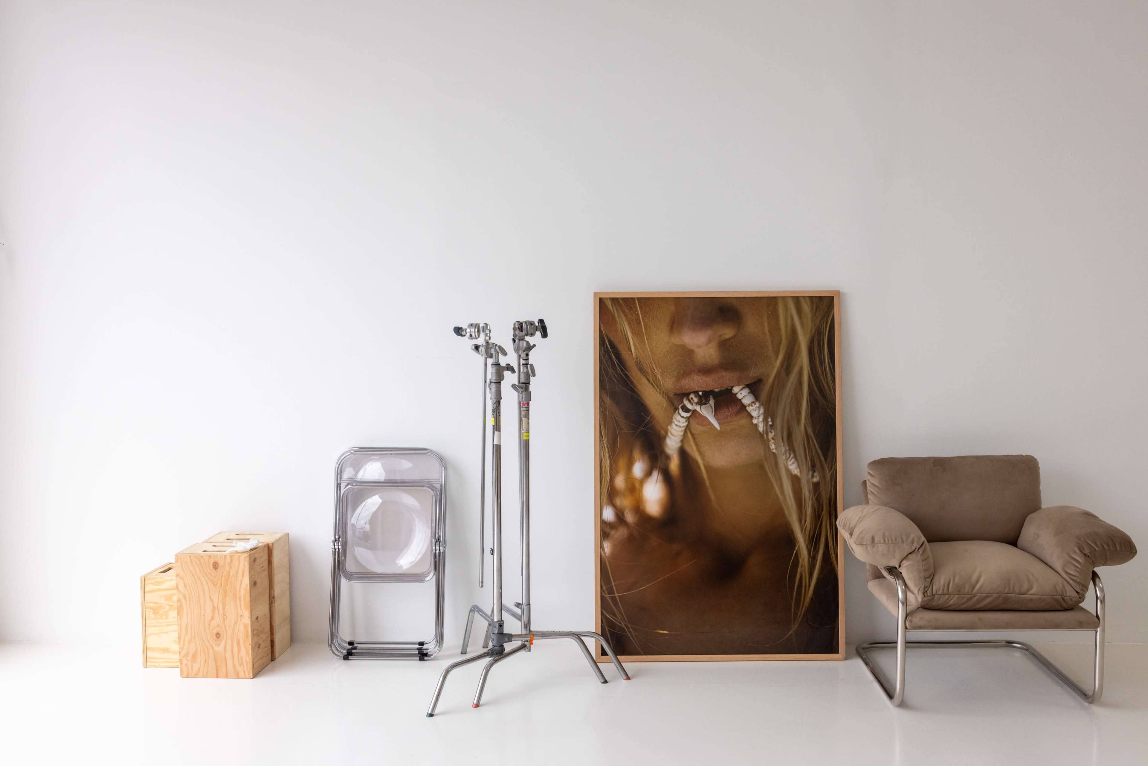 The image shows an interior space with a large framed artwork featuring a close-up of a person's face, alongside a beige chair, a folding chair, and a tripod.