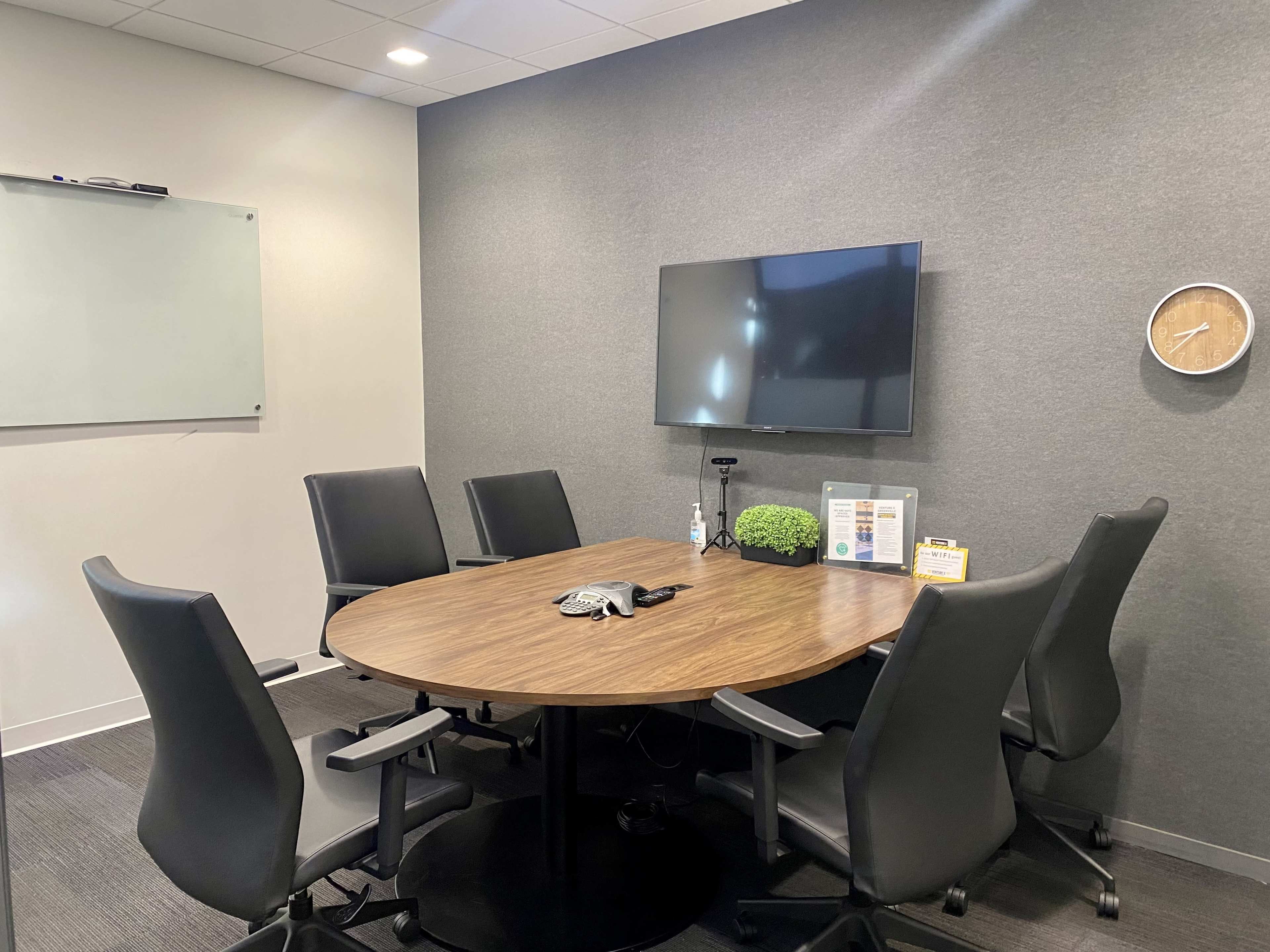 A conference room features a round wooden table surrounded by six black chairs, a wall-mounted television, a clock, and a whiteboard.