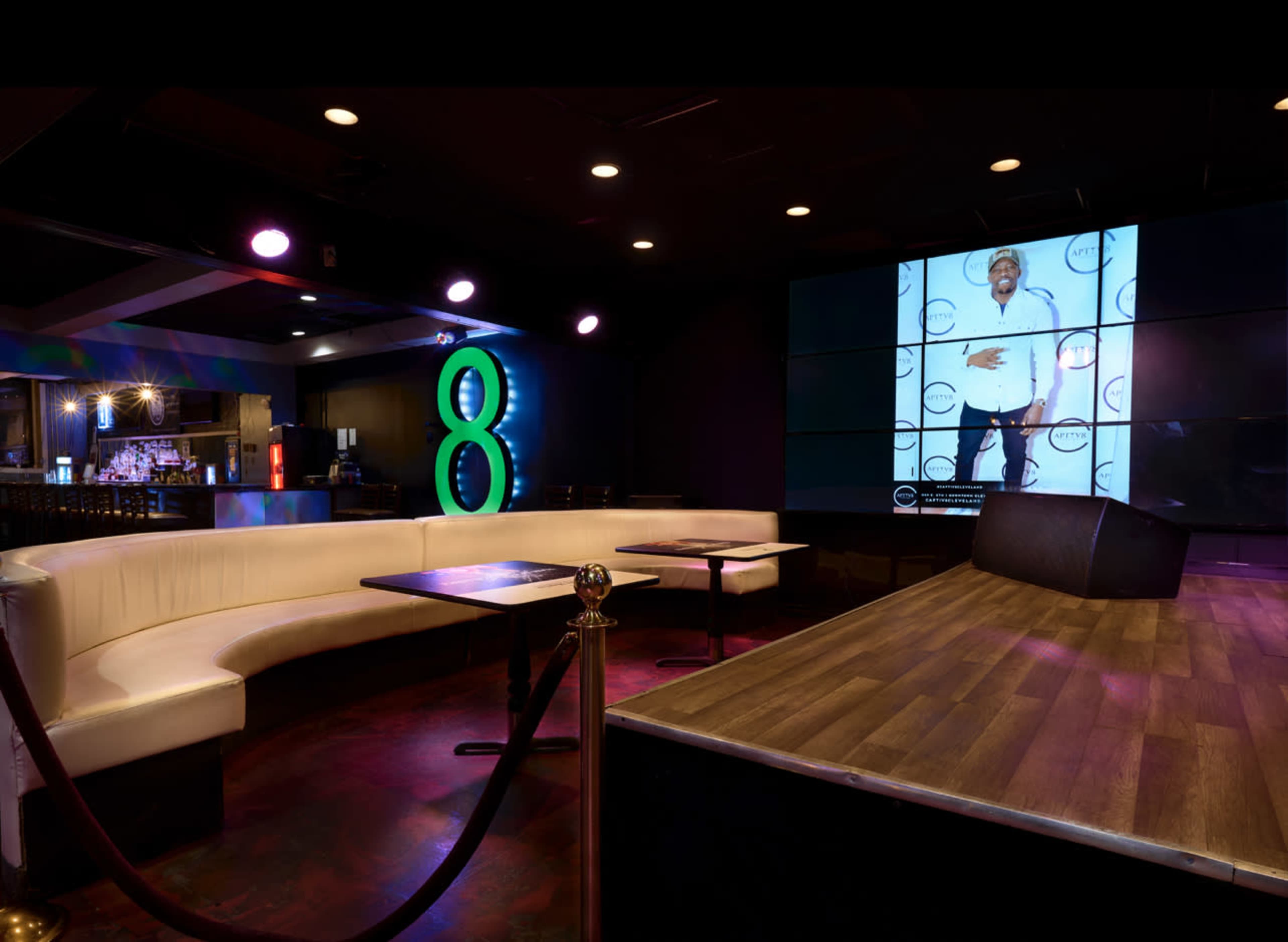 A dimly lit lounge features a curved white seating area, a bar in the background, and a large screen displaying a photo of a man.