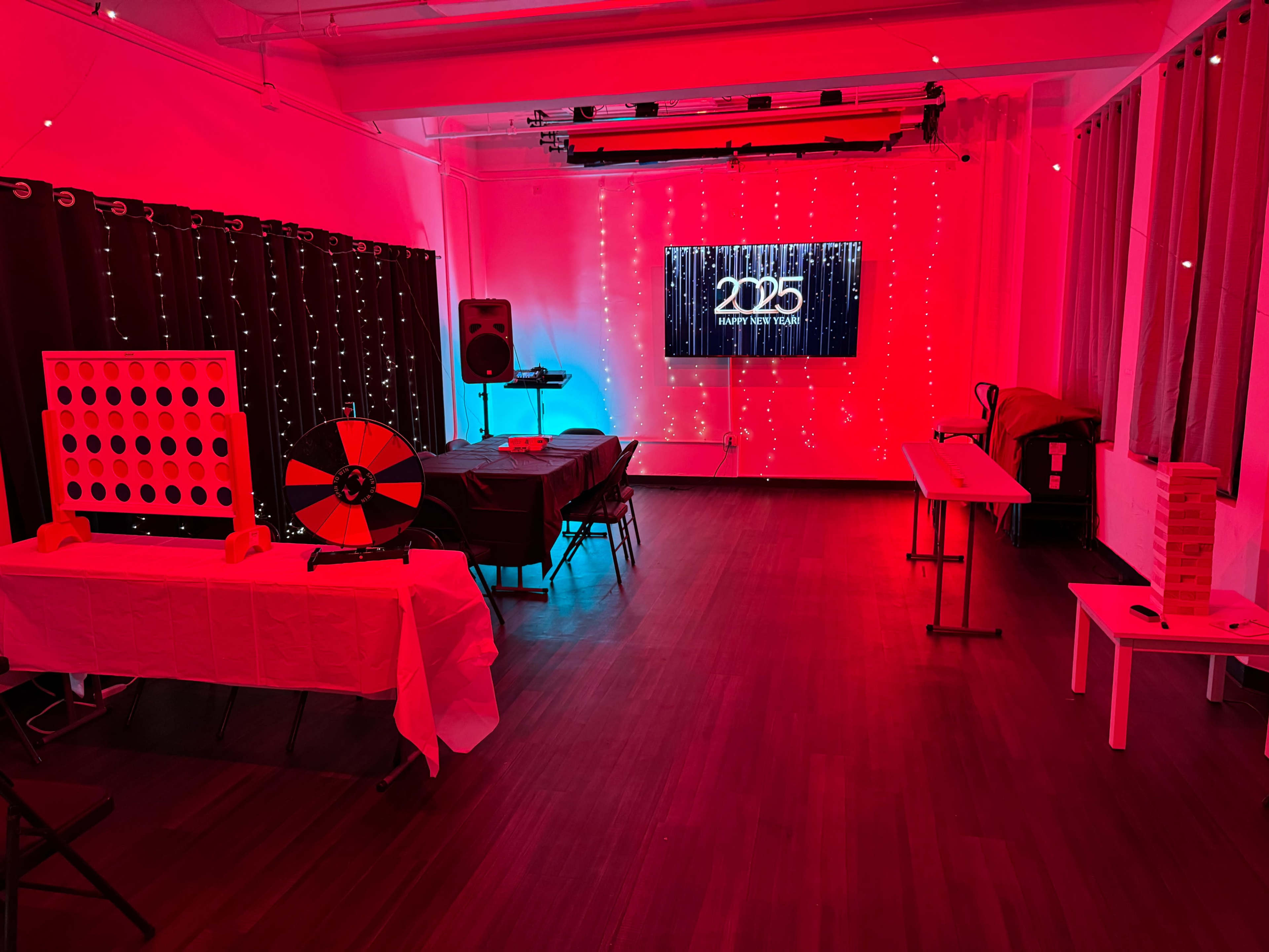 A brightly lit indoor space features a large screen showing "2025," a table set up for games, and red lighting with decorative string lights.