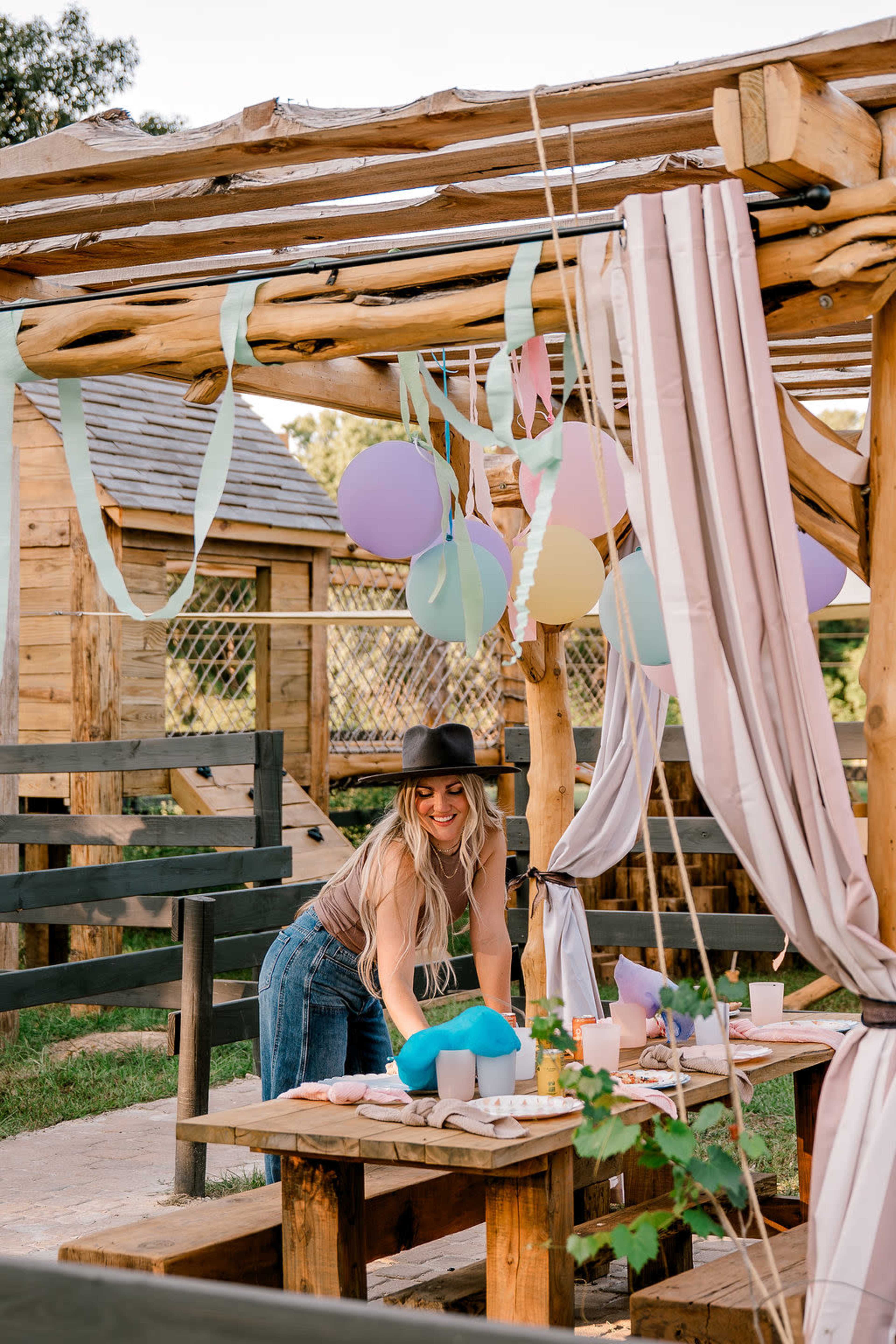 A woman in a hat is arranging decorations and food on a table under a wooden pergola adorned with colorful balloons and streamers at an outdoor setting.