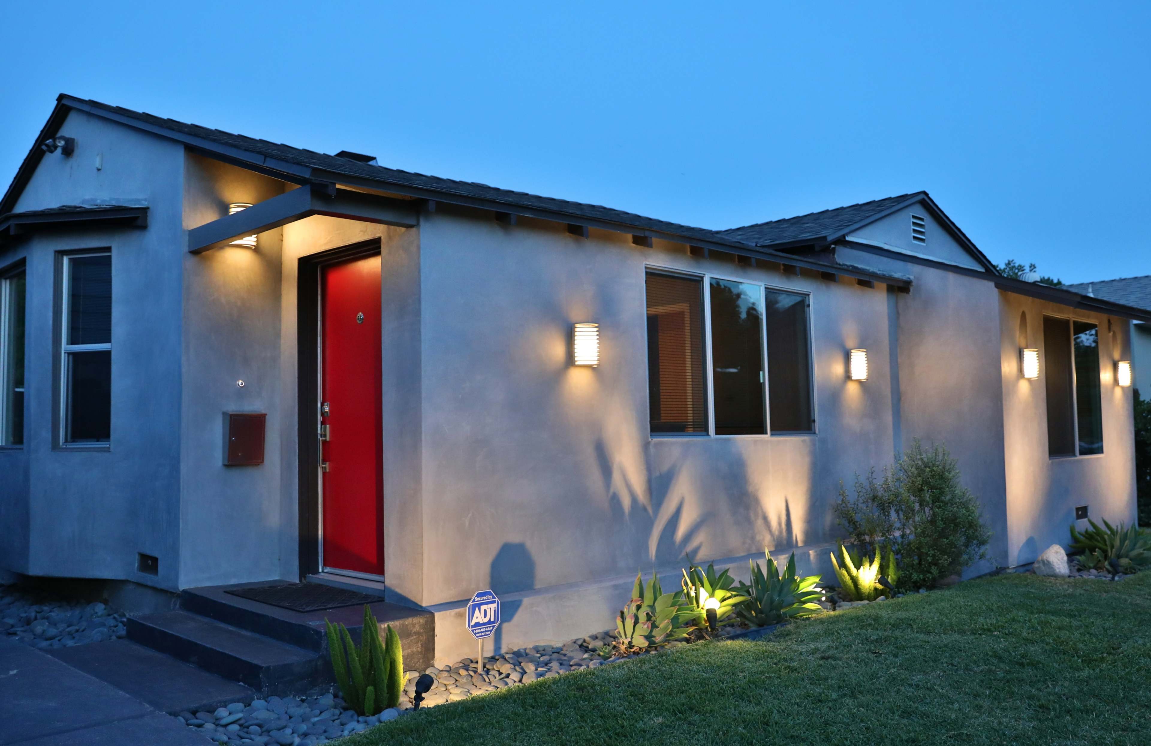 The image shows a modern house with a red front door, illuminated by exterior lights, and surrounded by a landscaped yard.