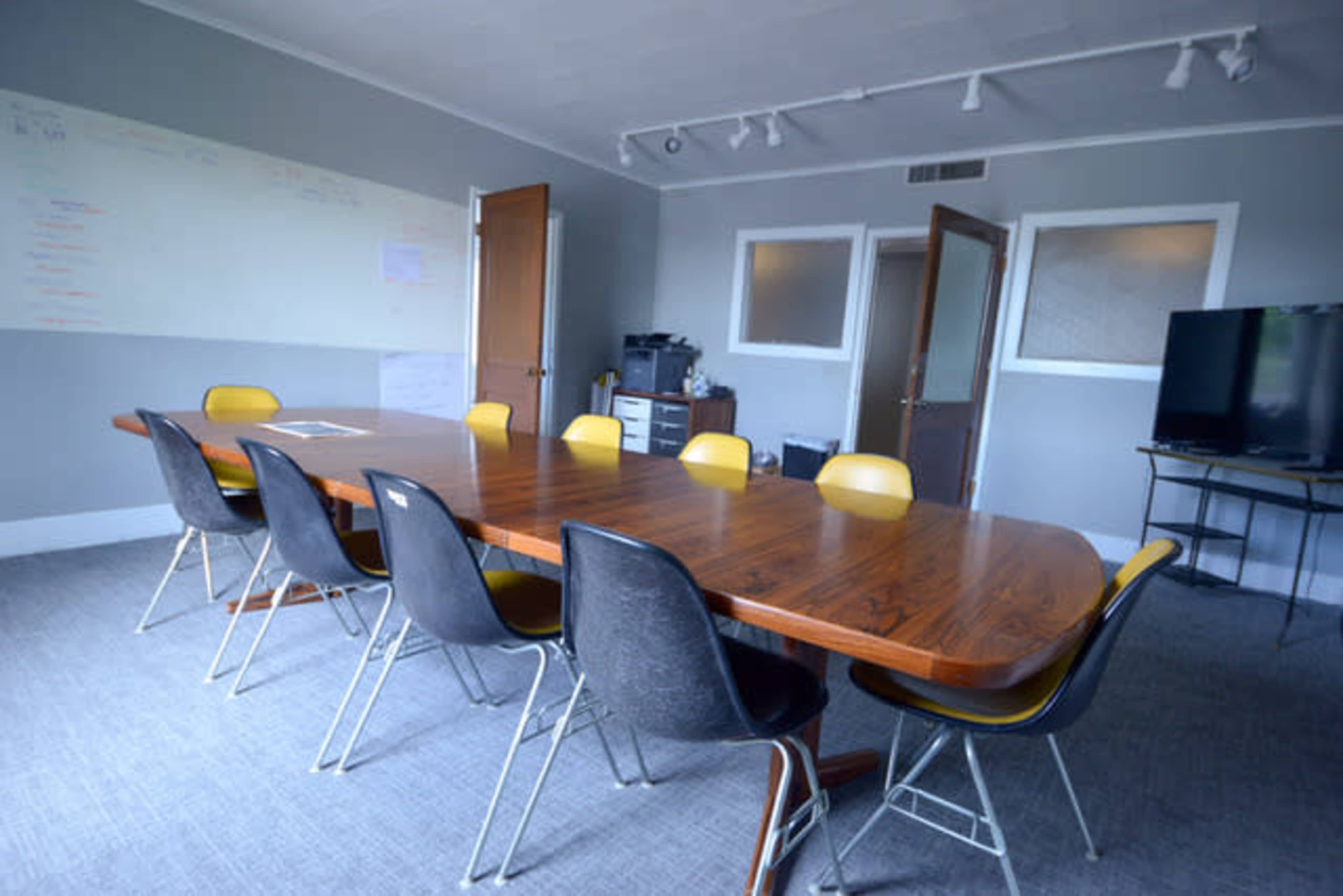 A long wooden conference table surrounded by black and yellow chairs is situated in a gray meeting room with a large TV and whiteboard.